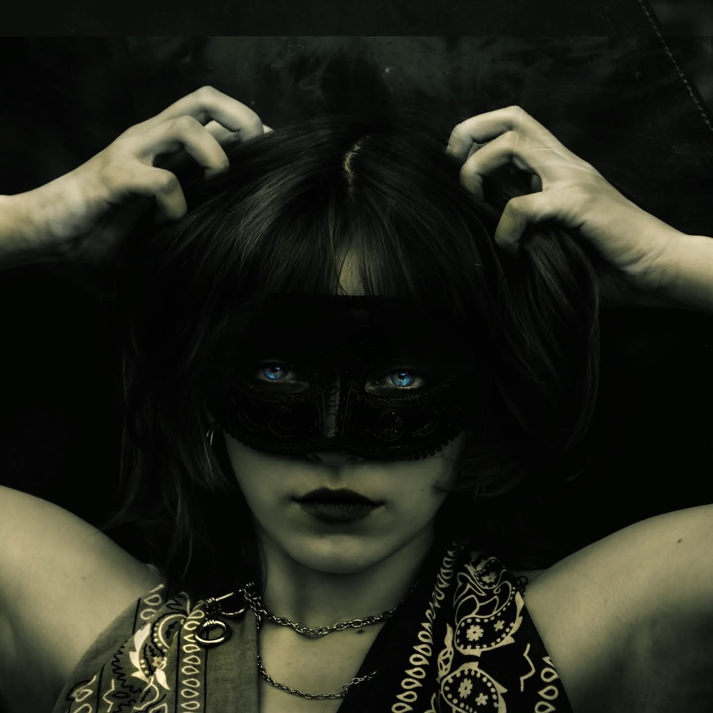 Portret kobiety w czarnej masce, z intensywnie niebieskimi oczami, dłońmi we włosach i wzorem paisley na bluzce, na ciemnym tle.