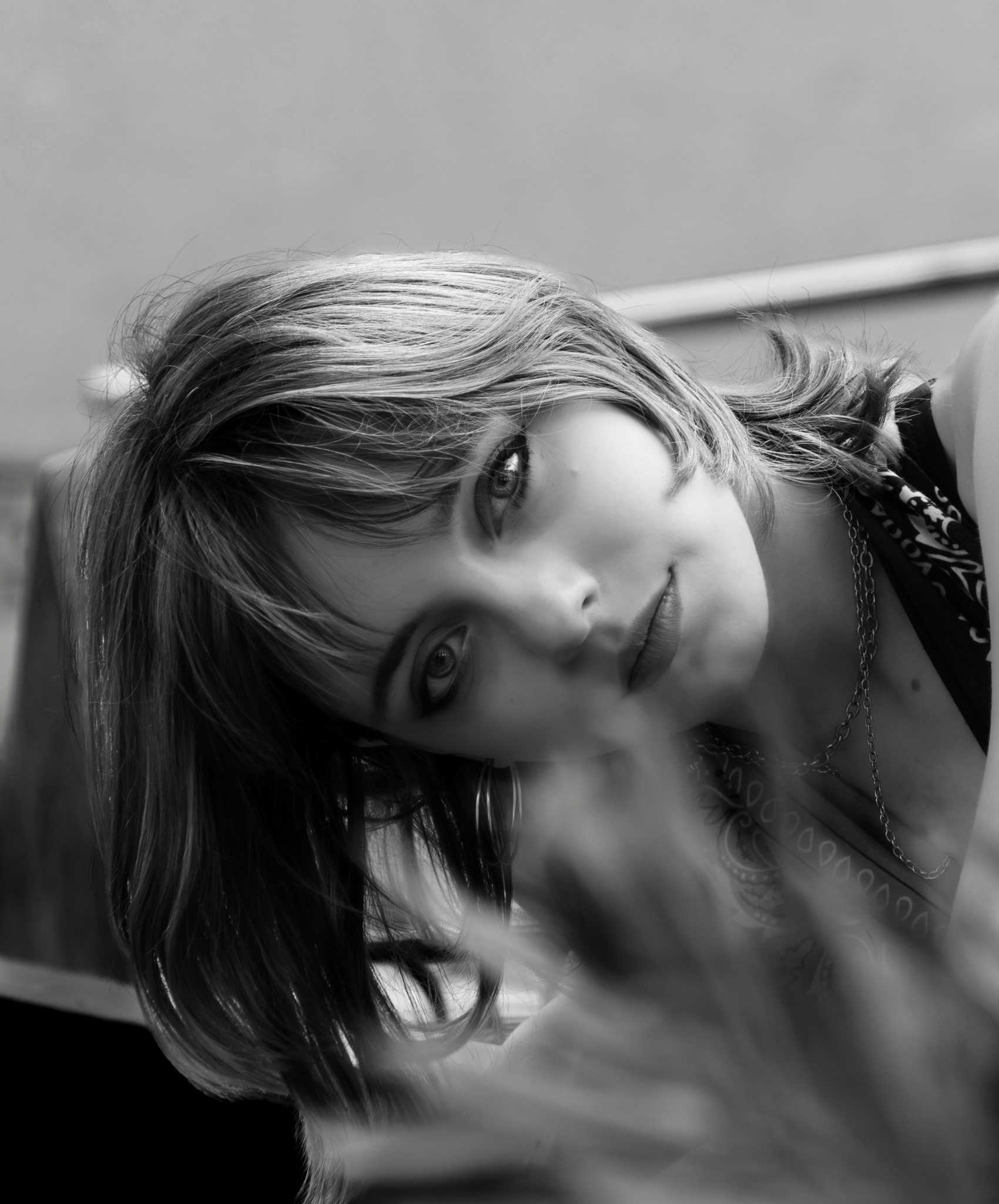 Czarno-białe, portretowe ujęcie kobiety z delikatnym uśmiechem, przechyloną głową i subtelnym makijażem oczu, z łańcuszkiem na szyi i wzorzystą chustą.