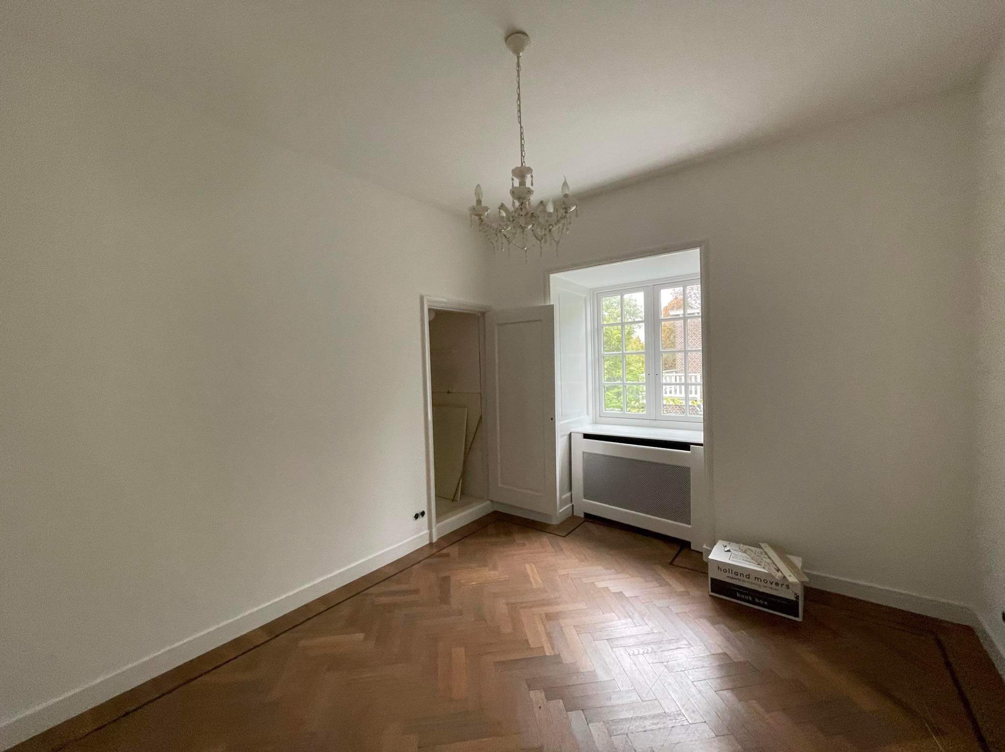 Puste pomieszczenie z parkietem ułożonym w jodełkę, świeżo pomalowane ściany na biało, kryształowy żyrandol i kartonowe pudła do przeprowadzki w rogu pokoju.