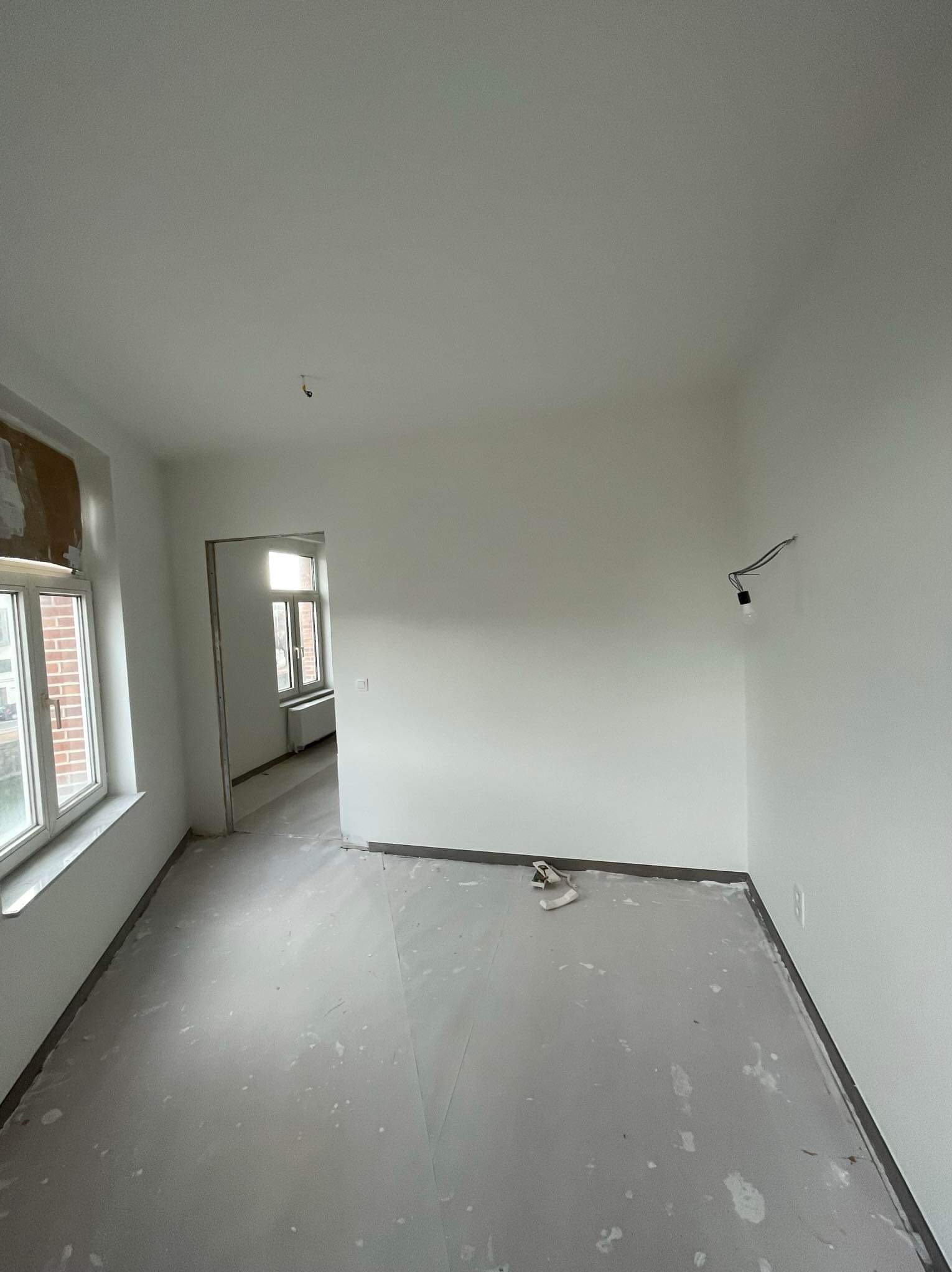 Pusty, świeżo odnowiony pokój z białymi ścianami, zabezpieczoną folią podłogą i wiszącą żarówką, widok na kolejne pomieszczenie przez otwarte drzwi.