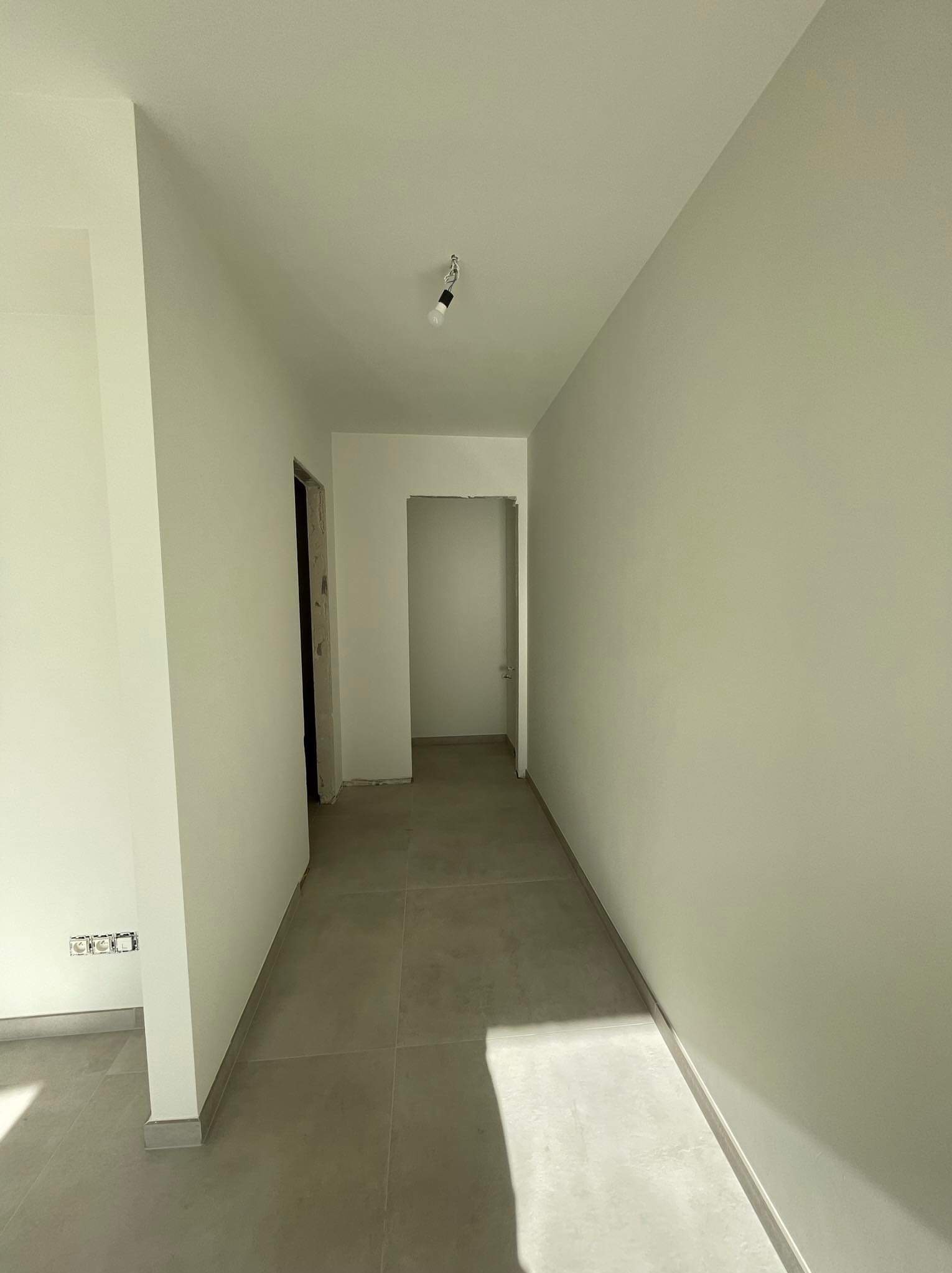Korytarz w jasnych barwach z szarą podłogą z płytek, świeżo po pracach wykończeniowych, widoczne gniazdka elektryczne i surowa żarówka.