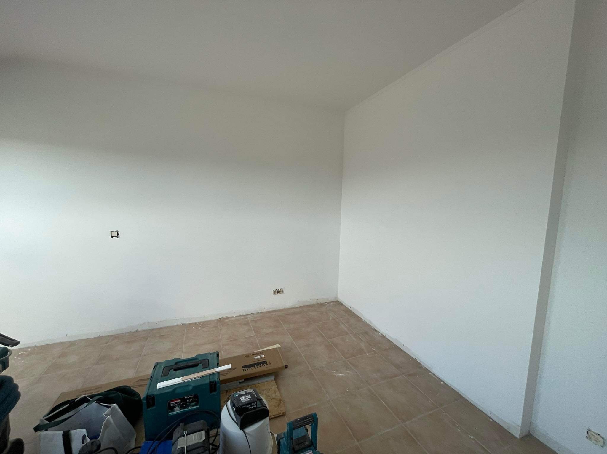 Przygotowane do malowania wnętrze z białymi ścianami, brązowymi płytkami podłogowymi, widocznymi narzędziami i akcesoriami malarskimi Makita w rogu pomieszczenia.