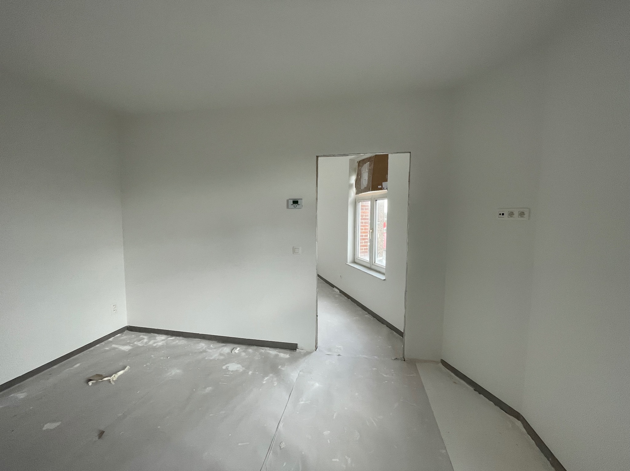 Pusty, świeżo pomalowany pokój z białymi ścianami i szarymi listwami przypodłogowymi; widoczne ślady po pracach na podłodze, przejście do kolejnego pomieszczenia z oknem.