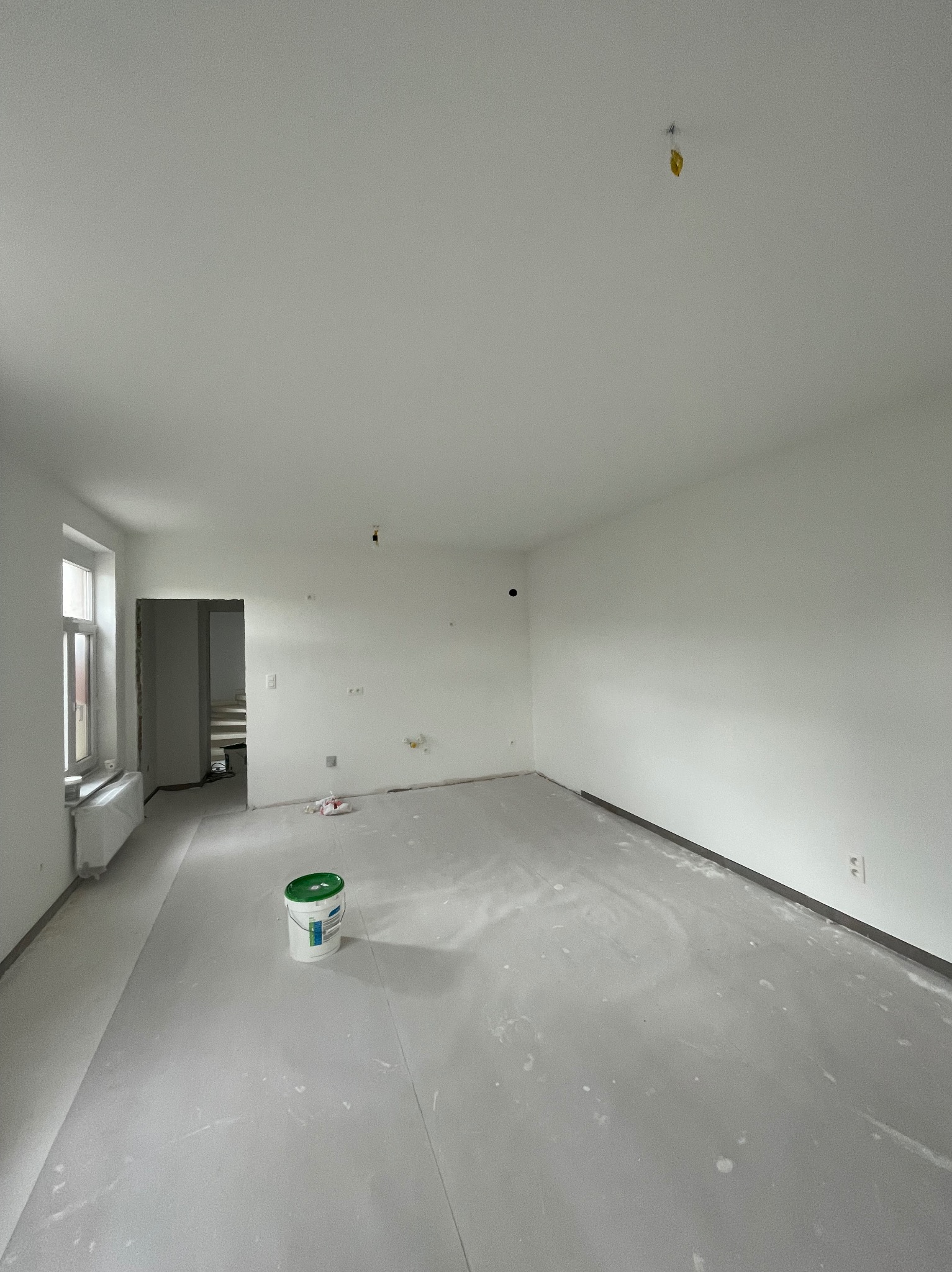Wnętrze w trakcie przygotowania do malowania: podłoga zabezpieczona folią, ściany świeżo zagruntowane, widoczne ślady po szpachlowaniu, wiadro z farbą w rogu pomieszczenia.