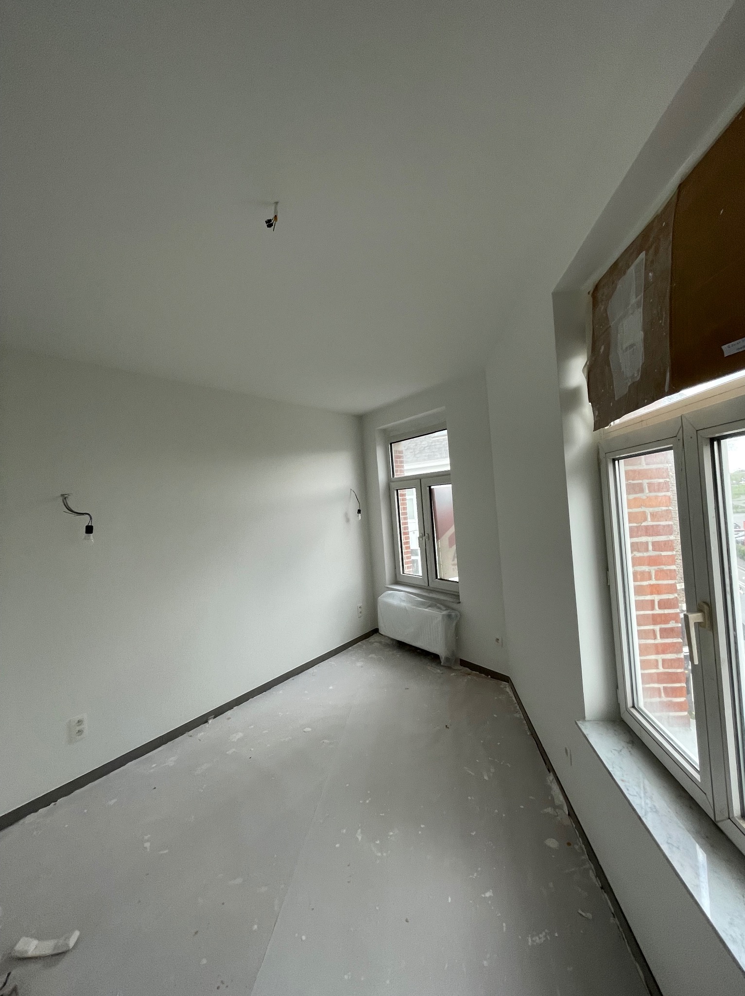 Wnętrze pokoju po świeżym pomalowaniu na biało, widoczne ślady farby na podłodze, okna z widokiem na ceglany budynek, przygotowane kable do oświetlenia.