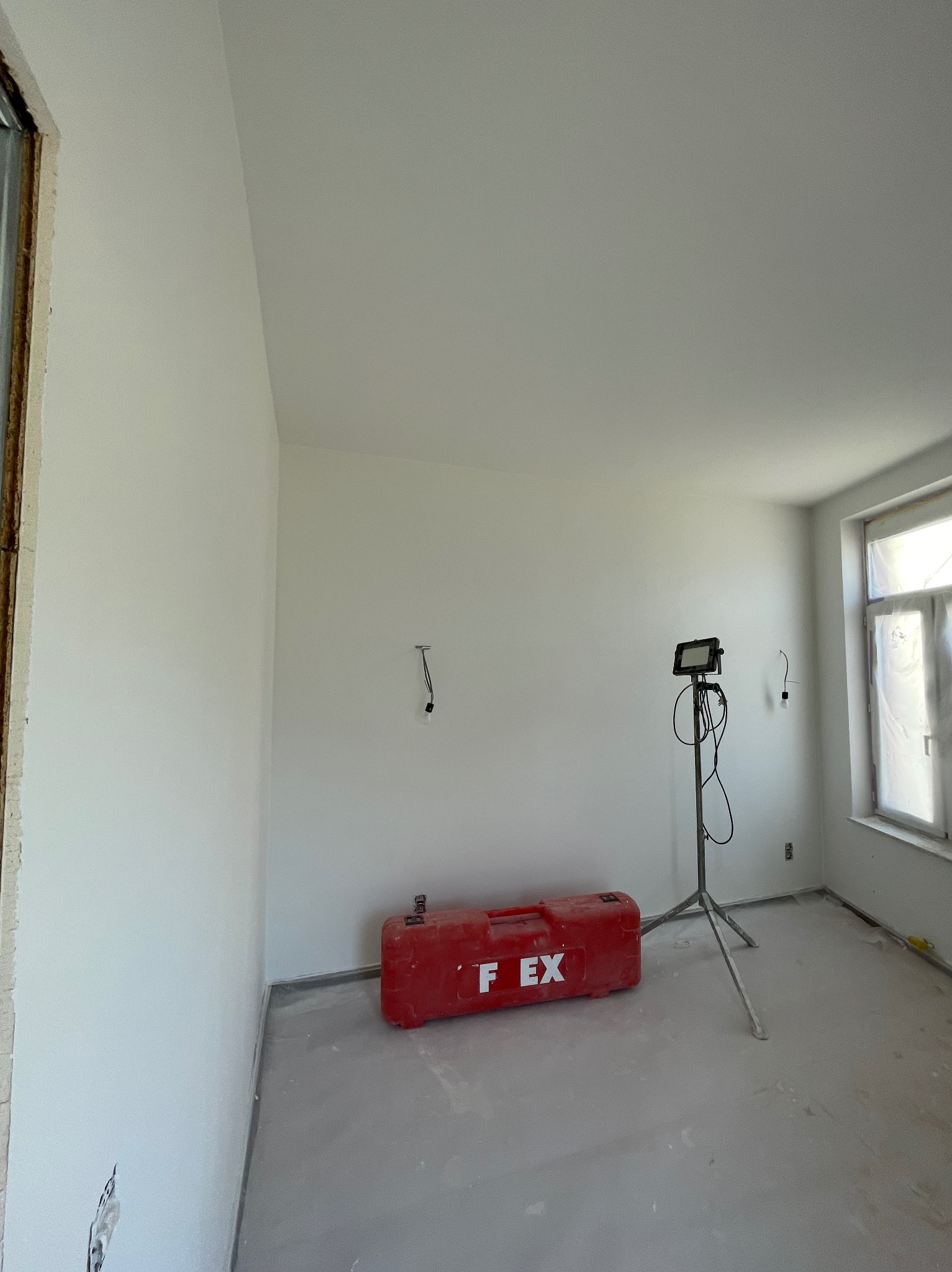 Przygotowane do odświeżenia pomieszczenie z białymi ścianami, widoczna czerwona skrzynka narzędziowa FLEX i lampa budowlana na trójnogu, okno zabezpieczone folią.
