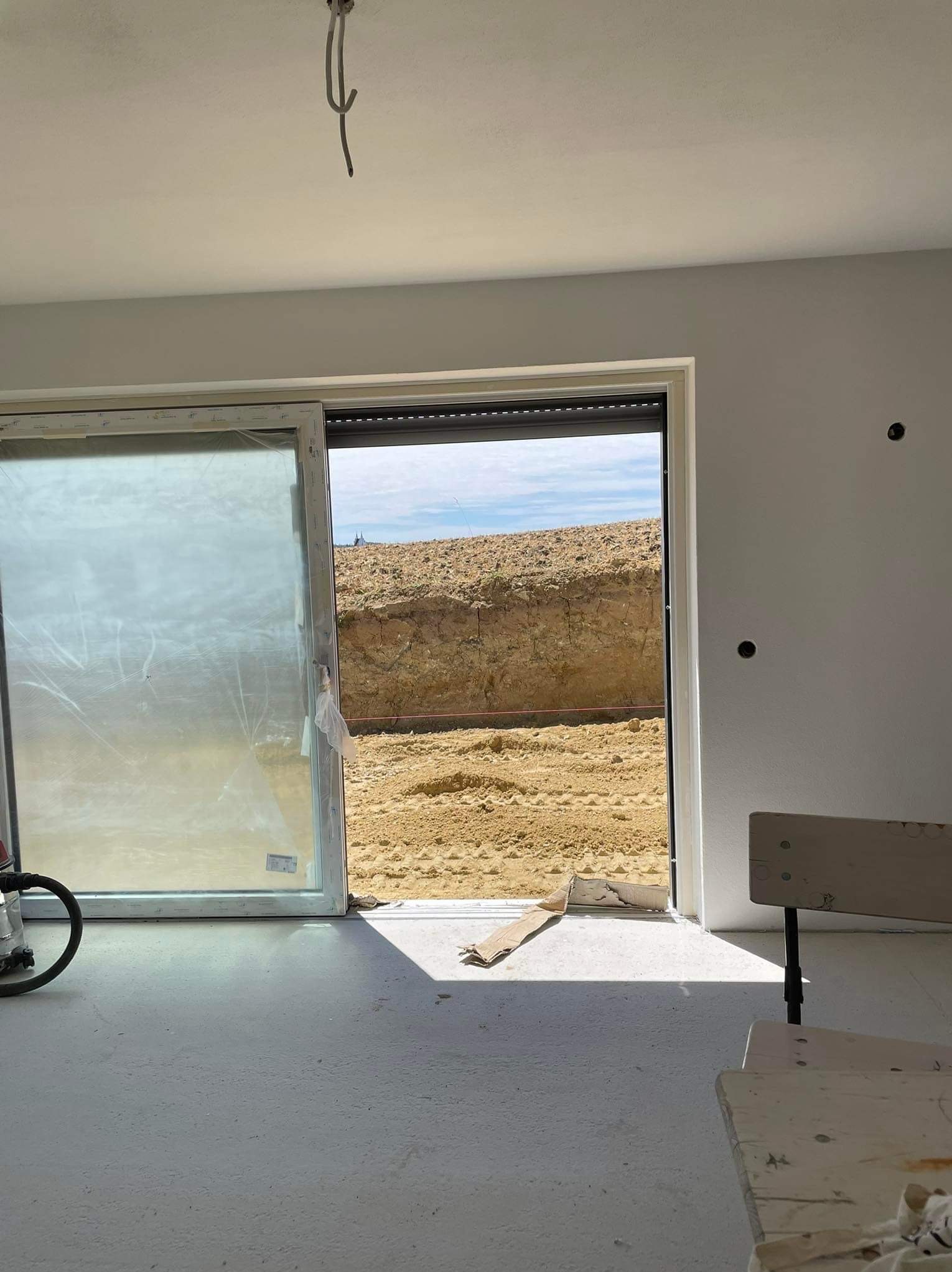 Wnętrze w trakcie prac wykończeniowych: surowe ściany, wylewka na podłodze, okno przesuwne z widokiem na wykop ziemny, widoczne kable ze ściany i sufitu.