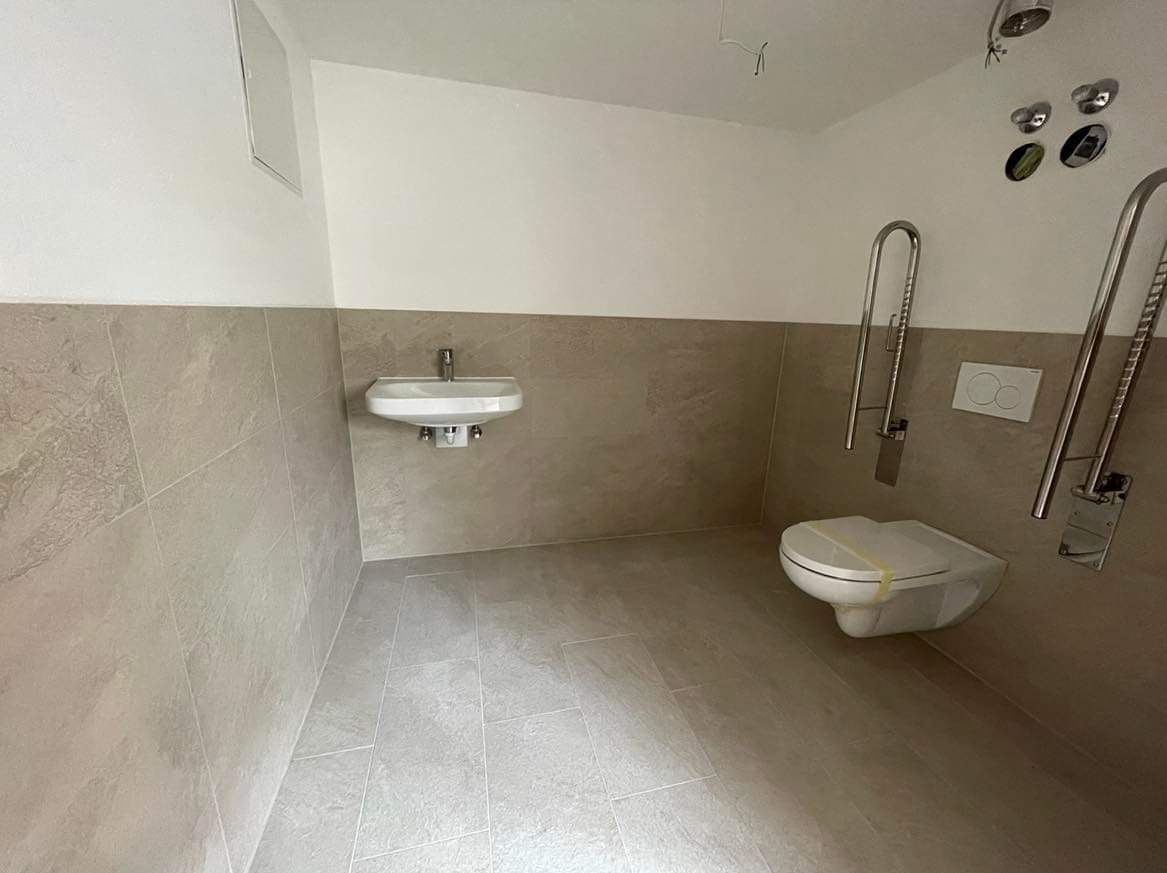 Wykończona łazienka w stonowanych beżach z wiszącą umywalką, toaletą z zabezpieczeniem transportowym i uchwytami dla osób niepełnosprawnych, widoczne niedokończone podłączenia oświetlenia na suficie.