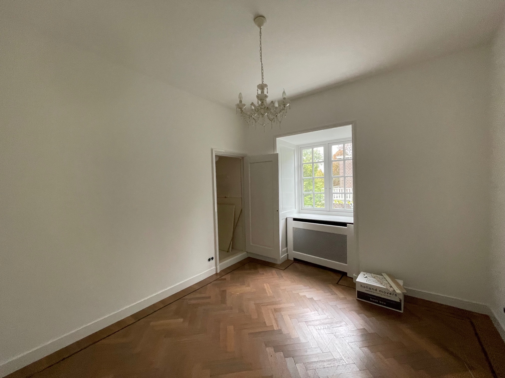 Pusty pokój z parkietem w jodełkę, białymi ścianami i kryształowym żyrandolem. Widoczne okno z widokiem na zieleń i kartonowe pudło firmy przeprowadzkowej.