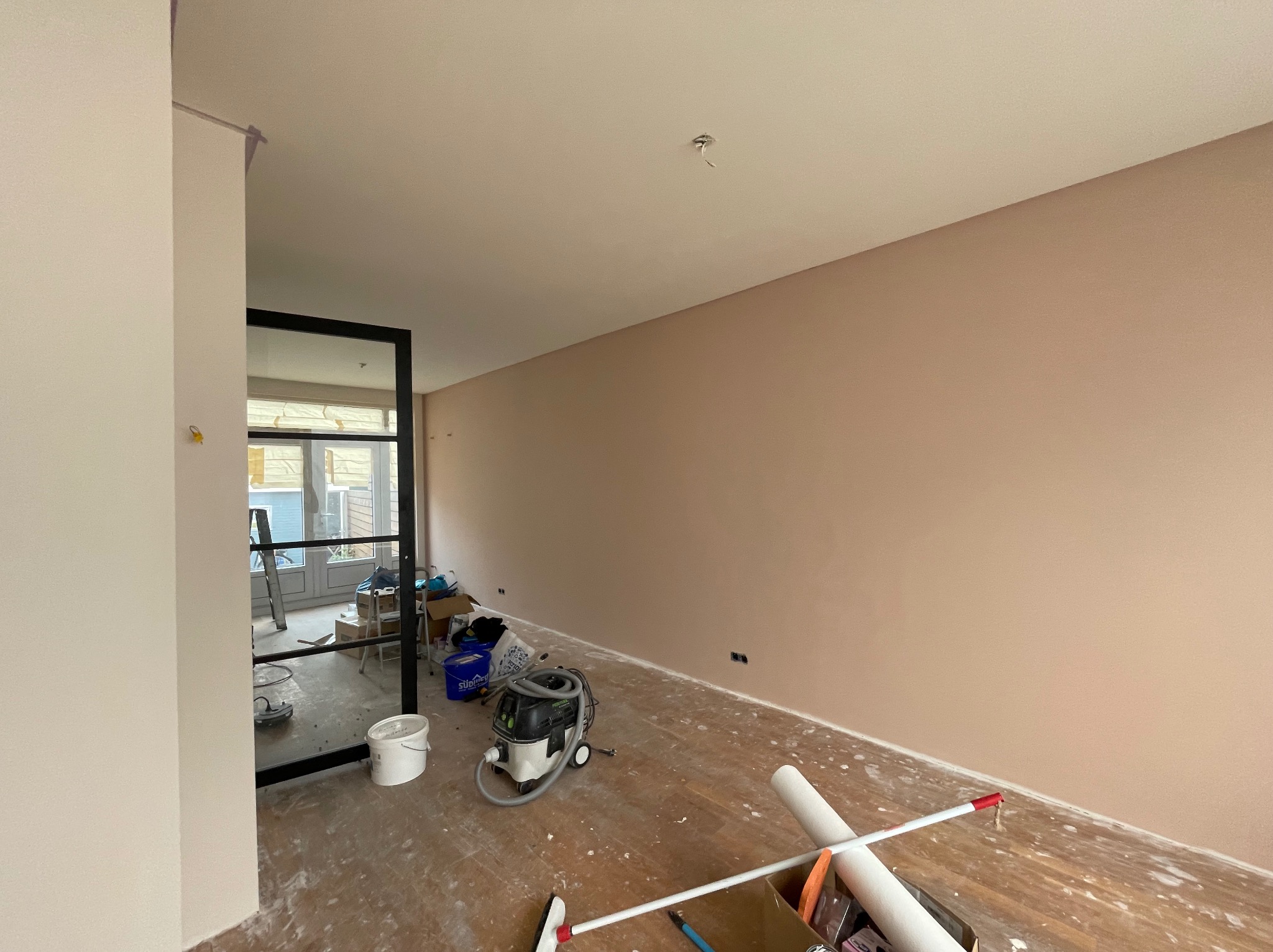 Pokój po malowaniu na delikatny róż, widoczne ślady farby na podłodze, narzędzia malarskie i odkurzacz przemysłowy.