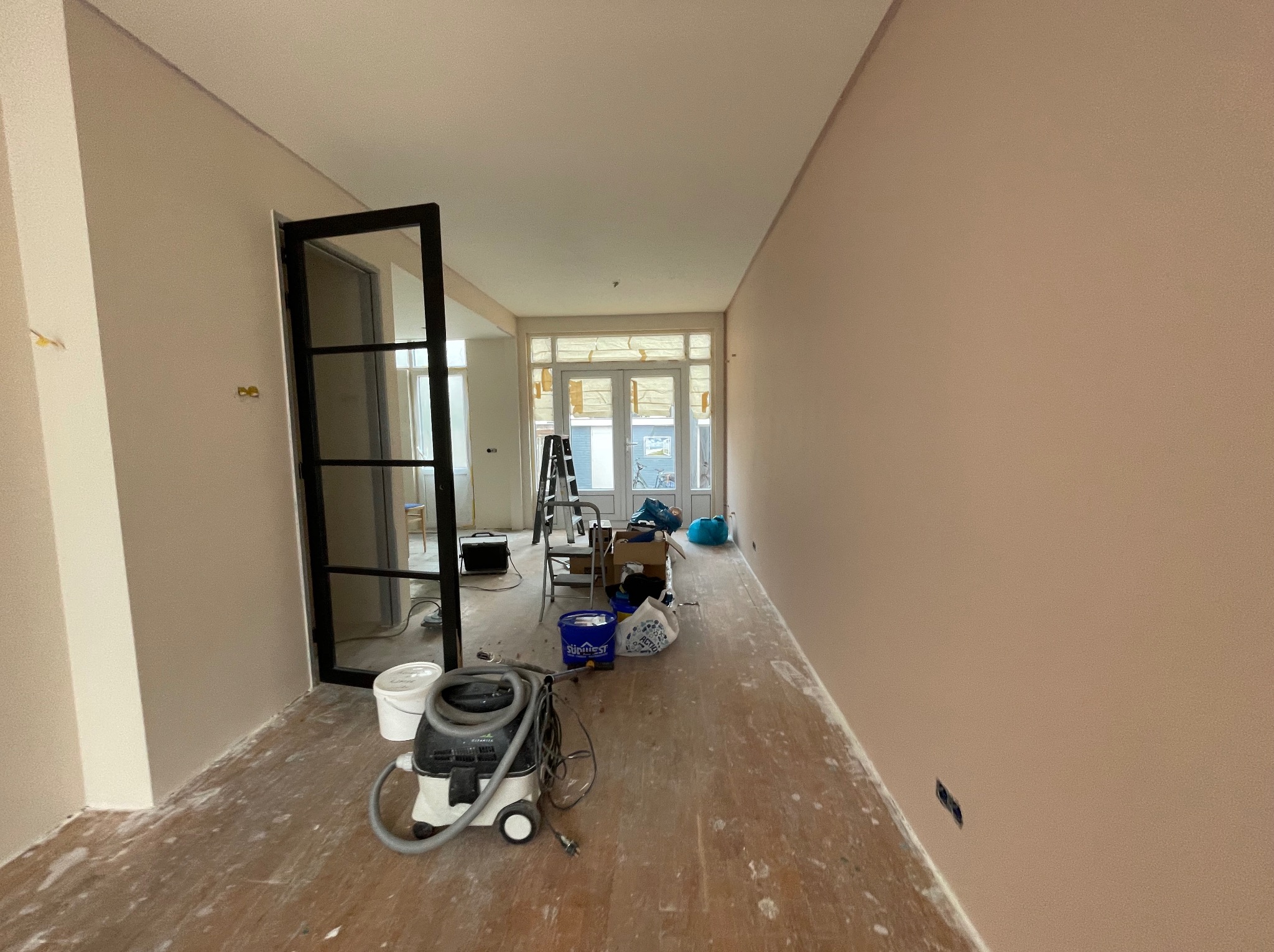 Pokój w trakcie odświeżania, ściany pomalowane na delikatny róż, widoczny odkurzacz przemysłowy, drabina i puszki z farbą, podłoga zabezpieczona folią malarską, czarna rama drzwiowa.