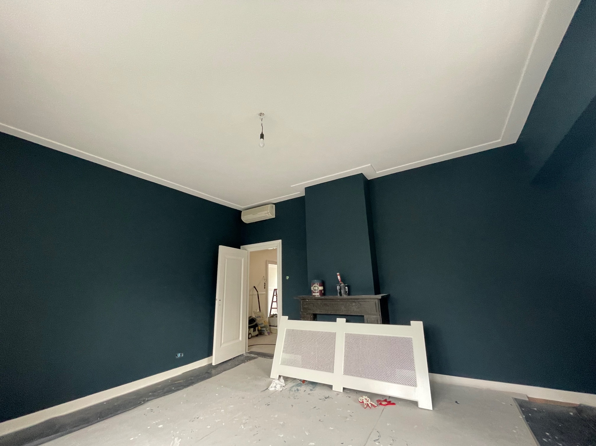 Pokój po świeżym pomalowaniu ścian na ciemny granat, z widocznymi śladami farby na zabezpieczonej podłodze i odsłoniętym grzejnikiem, gotowym do montażu.