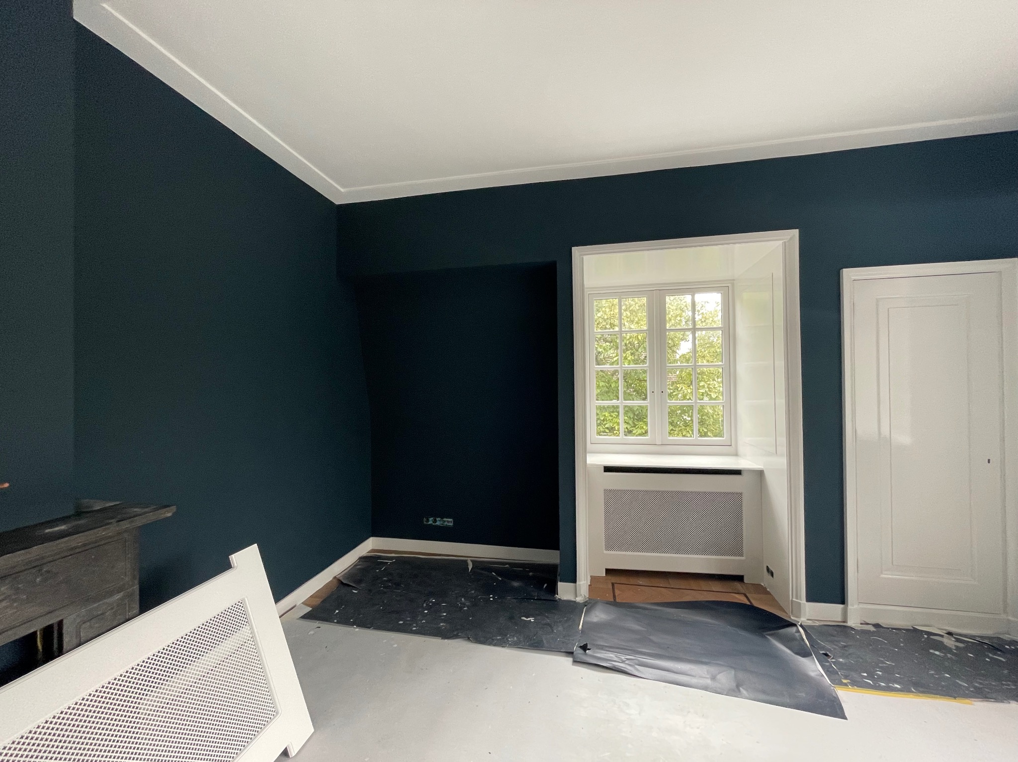 Pokój z ciemnymi, granatowymi ścianami i białym sufitem, w trakcie prac wykończeniowych, z zabezpieczoną folią podłogą i odstawionym grzejnikiem.