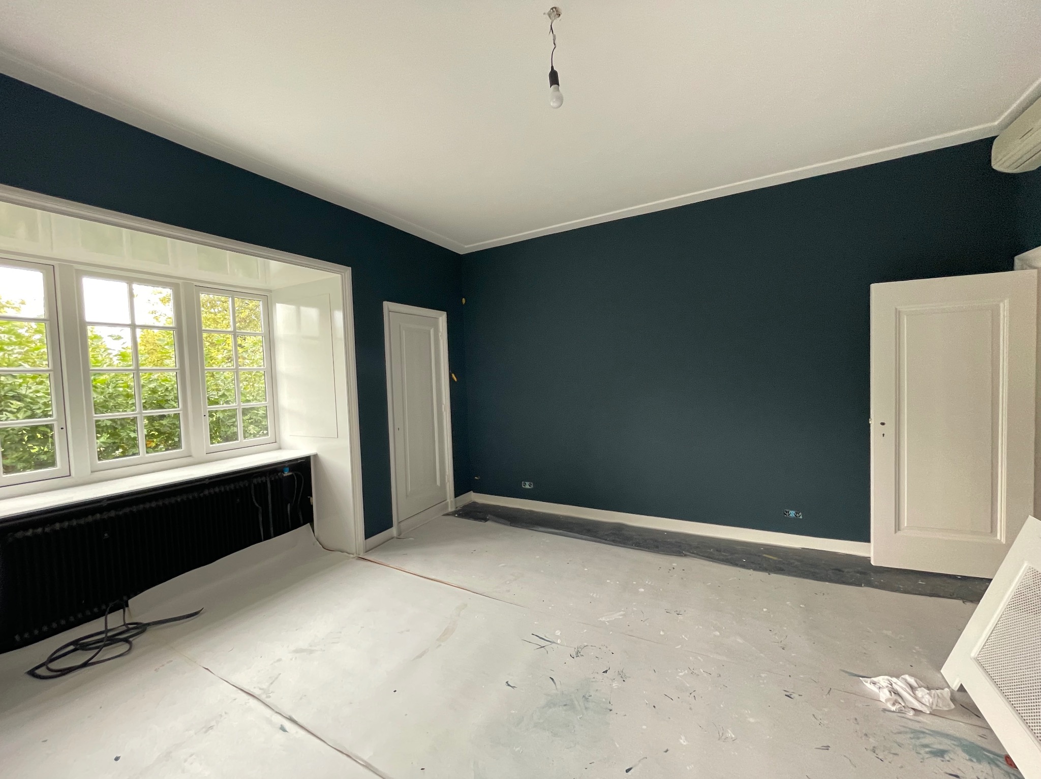 Pokój po pomalowaniu ścian na ciemny granat, widoczne zabezpieczone podłogi, białe okno z grzejnikiem i białe drzwi.