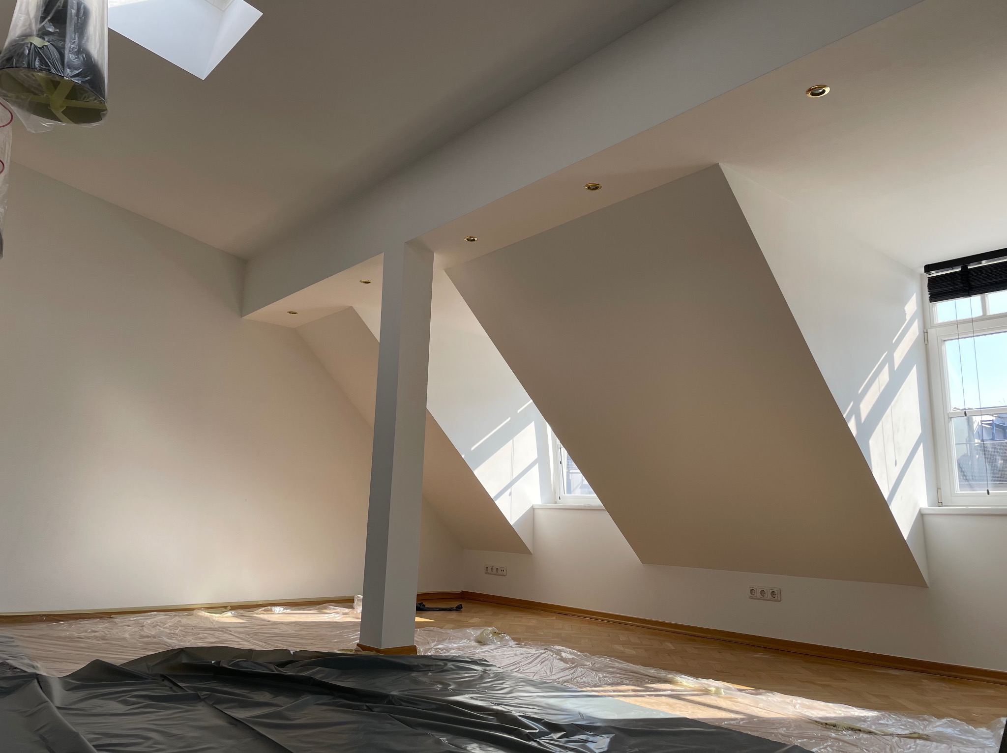 Jasne poddasze po świeżym pomalowaniu na biało, zabezpieczone folią podłogi, widoczne okno dachowe i okno w ścianie szczytowej.