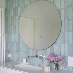 Plushy Pracownia Projektowa - Nowoczesna łazienka z turkusowymi, pionowo ułożonymi płytkami, okrągłym lustrem, białą umywalką nablatową, chromowaną armaturą, bukietem bzów w szklanym wazonie i świecą w kształcie kostki.