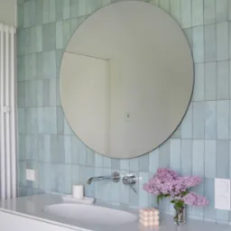 Nowoczesna łazienka z turkusowymi, pionowo ułożonymi płytkami, okrągłym lustrem, białą umywalką nablatową, chromowaną armaturą, bukietem bz&oacute;w w szklanym wazonie i świecą w kształcie kostki.