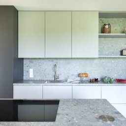 Plushy Pracownia Projektowa - Nowoczesna kuchnia w odcieniach szarości i bieli z blatem z lastryko, wyposażona w płytę indukcyjną, z otwartymi półkami z dekoracjami i produktami spożywczymi.