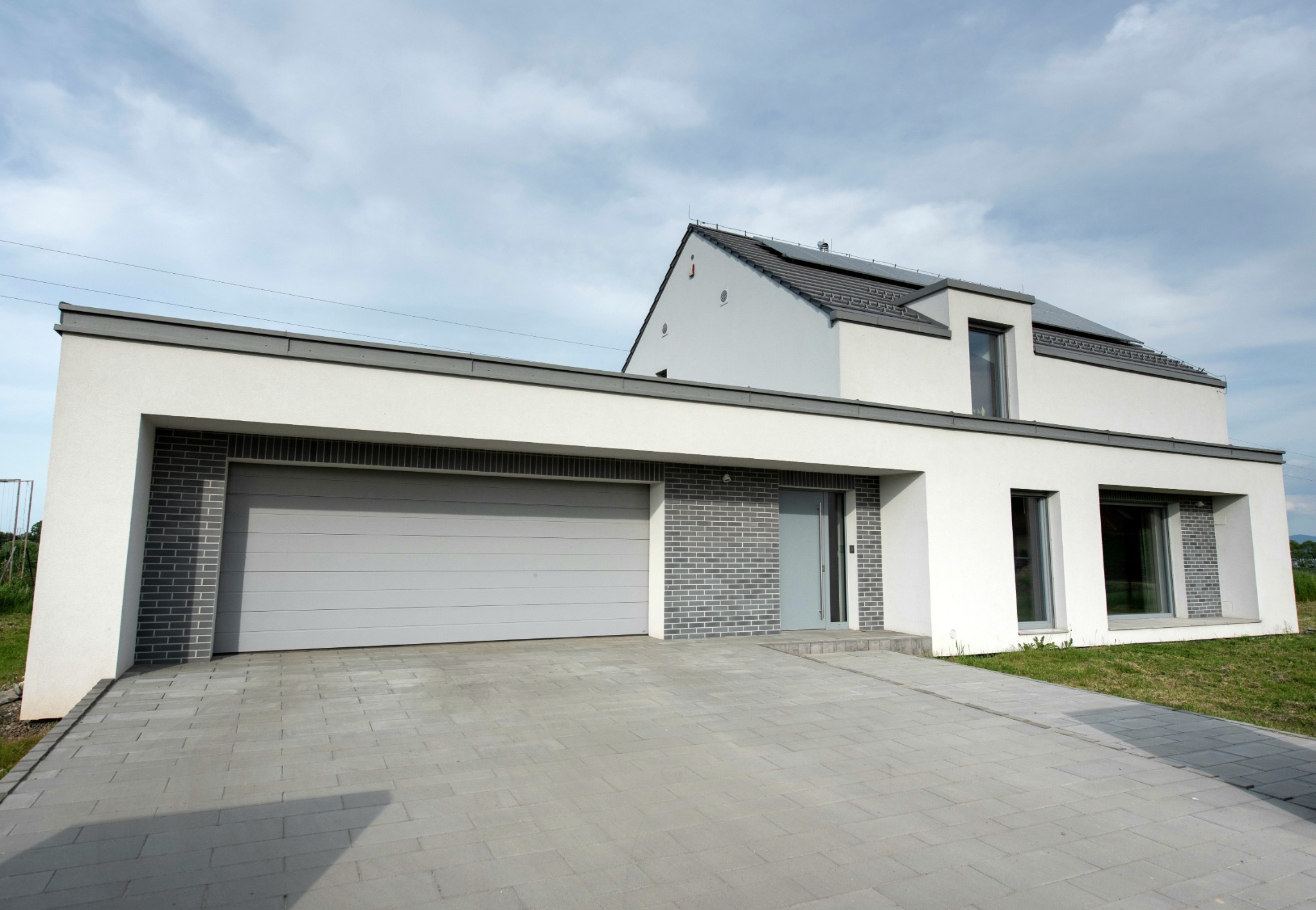 Nowoczesny, dwukondygnacyjny dom z płaskim dachem nad garażem i elewacją w kolorze białym i szarym, z szarą kostką brukową na podjeździe.