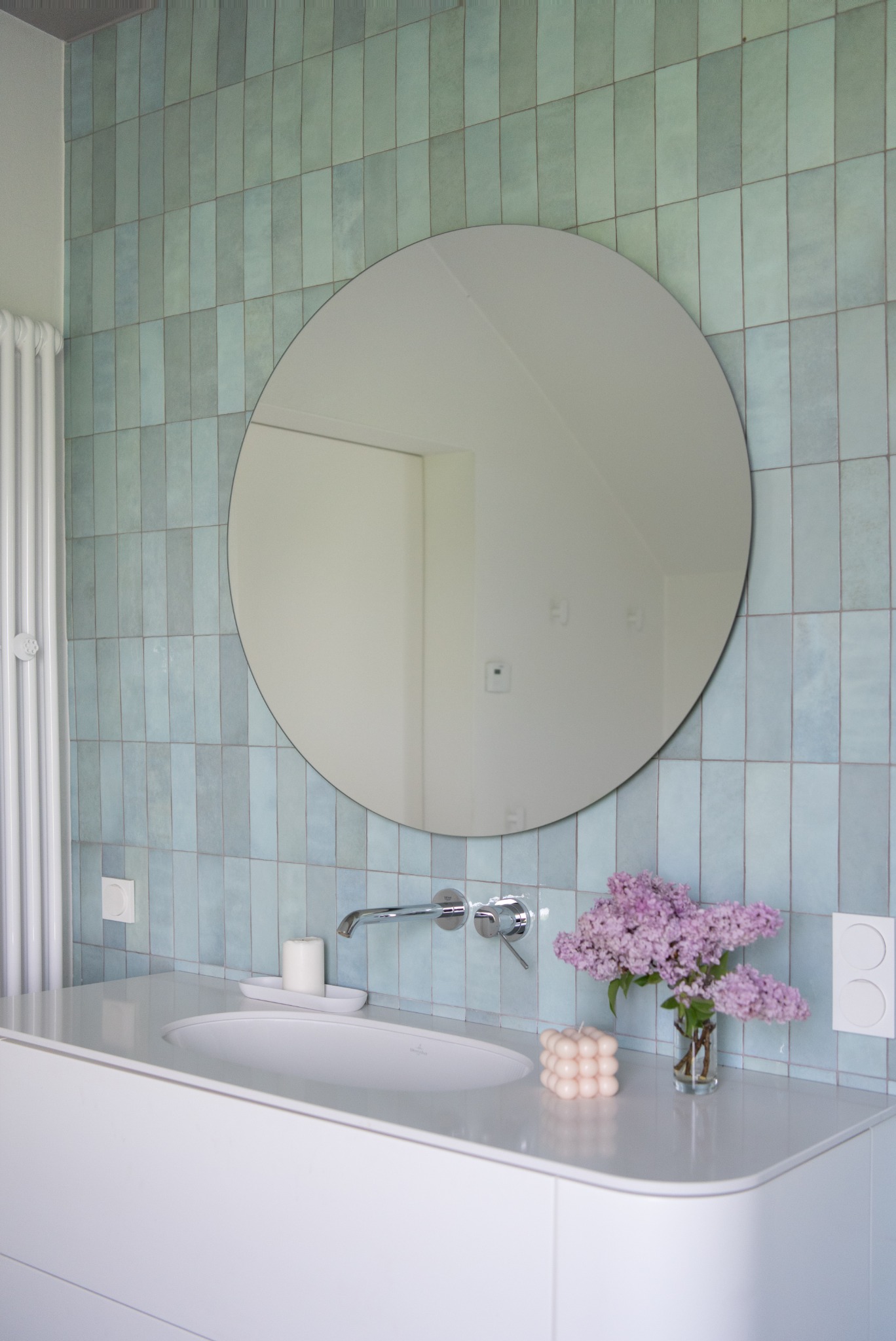 Nowoczesna łazienka z turkusowymi, pionowo ułożonymi płytkami, okrągłym lustrem, białą umywalką nablatową, chromowaną armaturą, bukietem bzów w szklanym wazonie i świecą w kształcie kostki.