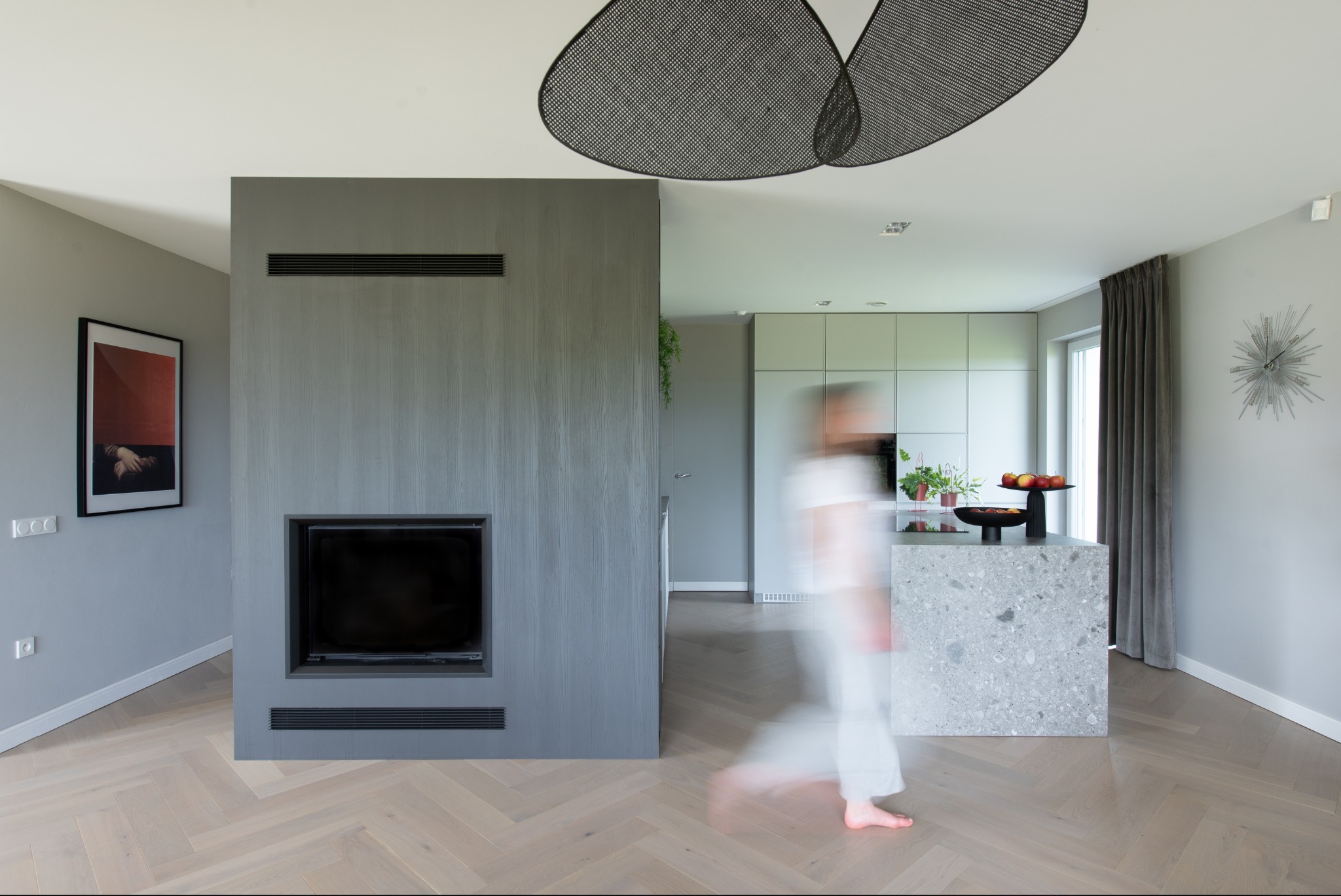 Nowoczesne wnętrze domu z szarą zabudową kominka, wyspą kuchenną z blatem z lastryko i parkietem w jodełkę, rozmyta sylwetka osoby w ruchu.