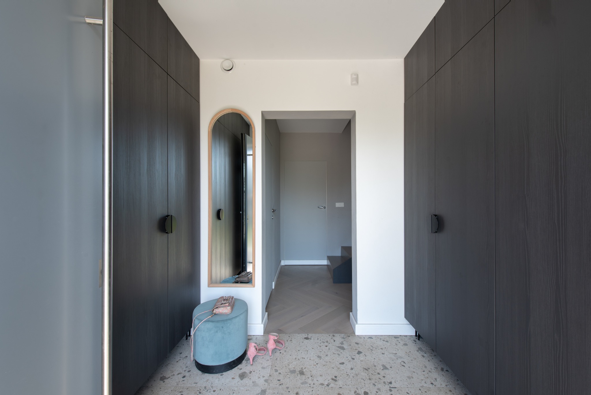 Nowoczesny przedpokój z zabudową szaf w kolorze grafitowym, okrągłym lustrem w drewnianej ramie, pufą w kolorze błękitnym oraz posadzką lastryko