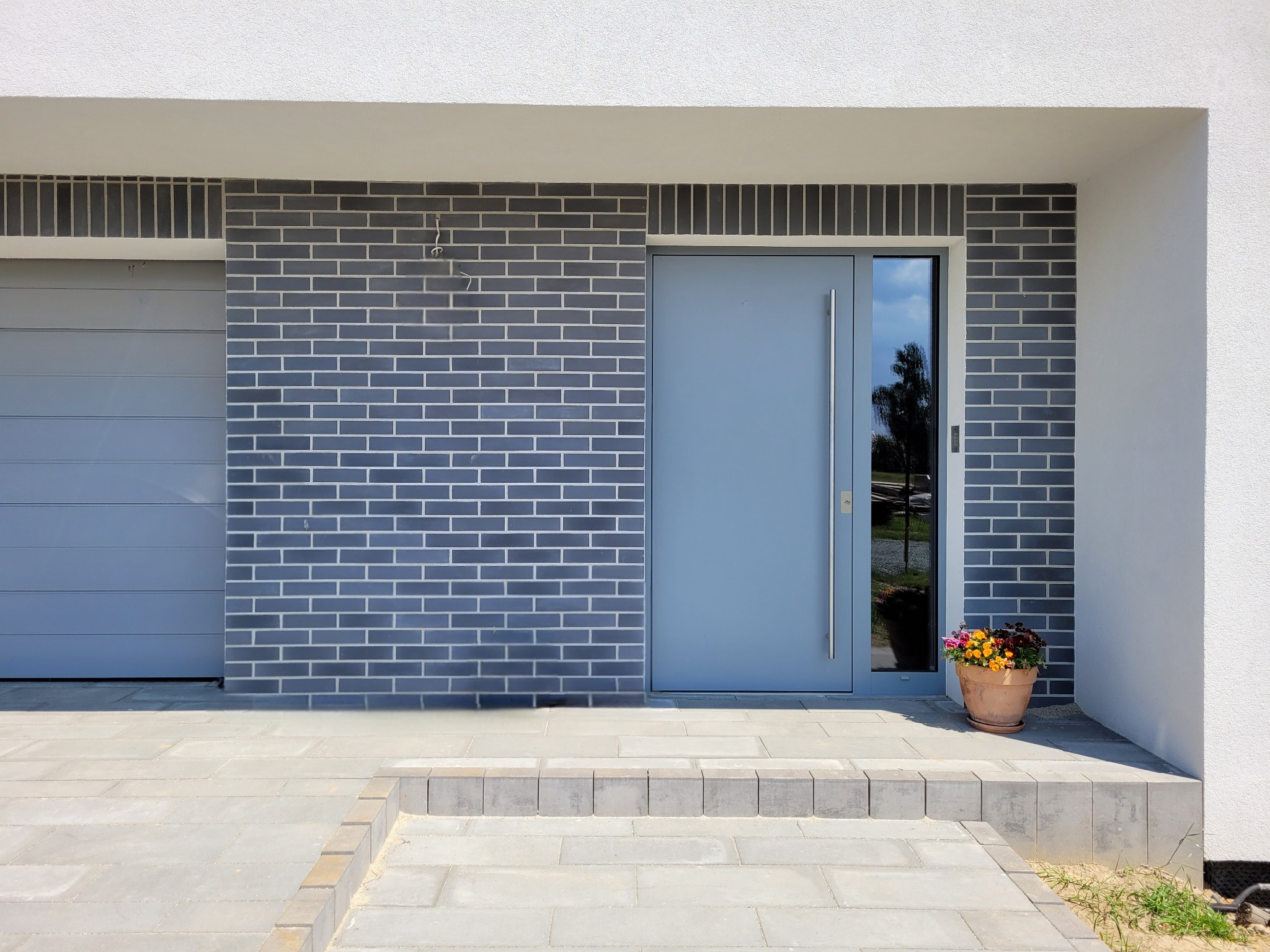 Nowoczesne wejście do domu z szarej cegły klinkierowej, szare drzwi z przeszkleniem, szara brama garażowa i donica z kolorowymi kwiatami.
