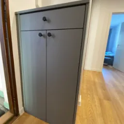 Szara szafka na buty z dwoma drzwiczkami i szufladą, czarne okrągłe uchwyty, Mierzyn. Widok na wnętrze mieszkania z drewnianą podłogą.