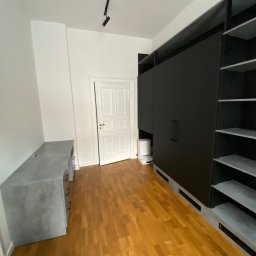 HS MEBLE Hubert Słowikowski - Czarna szafa na wymiar z półkami, szary blat biurka i parkiet w pokoju. Widoczny odkurzacz automatyczny przy szafie. Minimalistyczny design wnętrza.