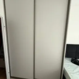 Dwudrzwiowa szafa przesuwna z matowego szkła i ramą w kolorze szaro-brązowym, widoczna w sypialni na tle łóżka z ciemnym zagłówkiem.