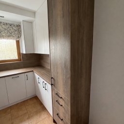 HS MEBLE Hubert Słowikowski - Nowoczesna kuchnia z białymi szafkami i wysoką, drewnopodobną zabudową. Uwagę zwracają czarne, minimalistyczne uchwyty i brązowa podłoga z płytek.