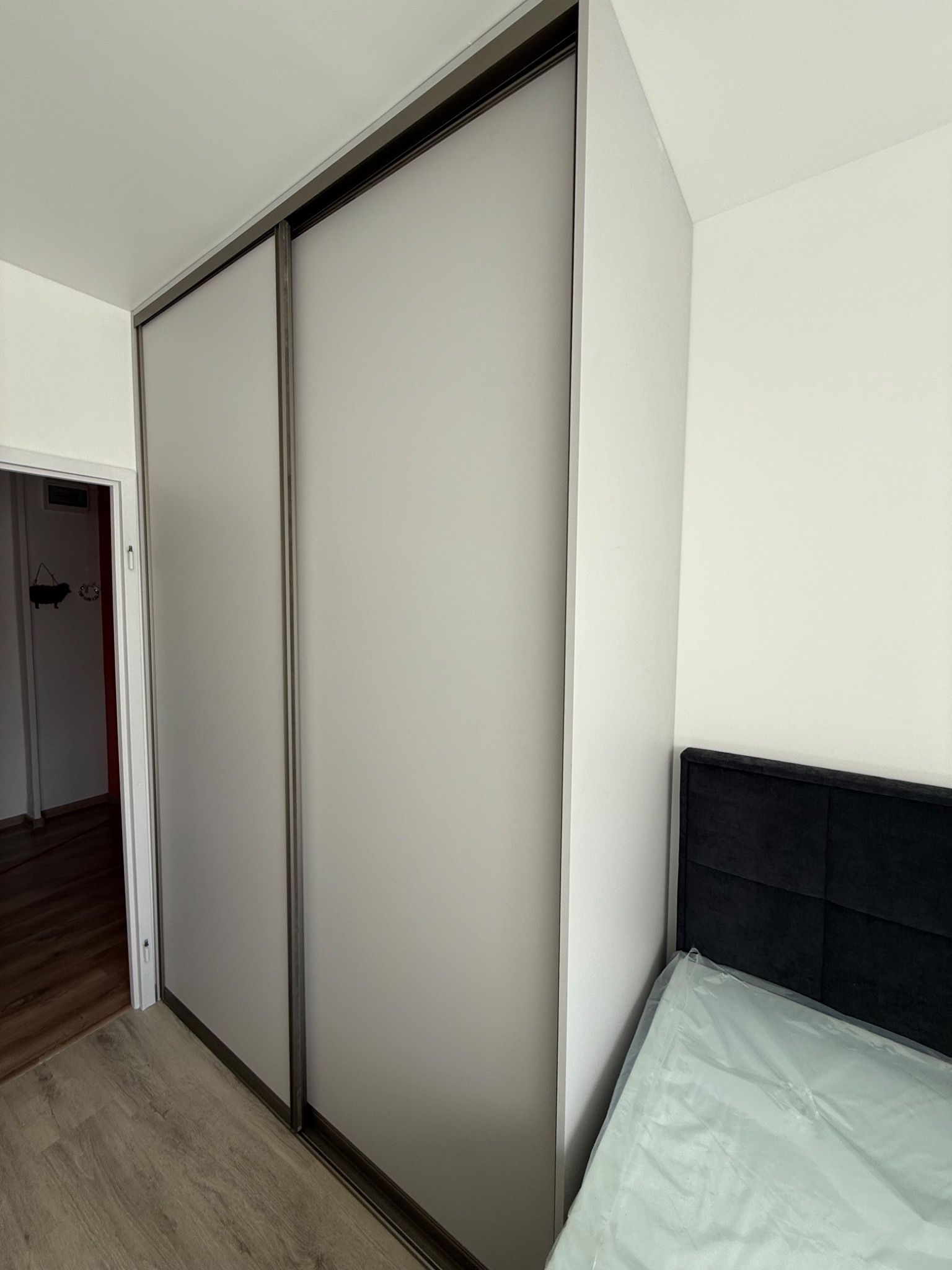 Dwudrzwiowa szafa wnękowa z przesuwnymi, matowymi drzwiami w pokoju z drewnianą podłogą i czarnym zagłówkiem łóżka. Widok z boku, drzwi zamknięte.