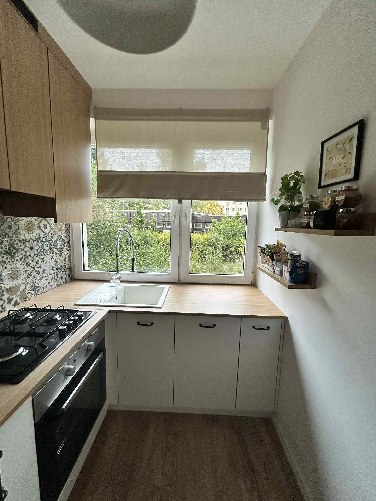 Jasna kuchnia z drewnianymi szafkami górnymi i białymi dolnymi, z blatem imitującym drewno. Nad zlewem roleta okienna i widok na zieleń. Dekoracyjne płytki nad kuchenką.