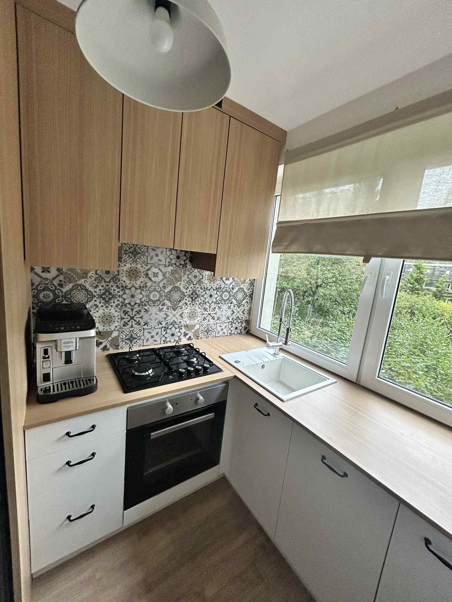 Nowoczesna, jasna kuchnia z drewnianymi szafkami, wzorzystą mozaiką nad blatem, widokiem na zieleń za oknem i ekspresem do kawy.