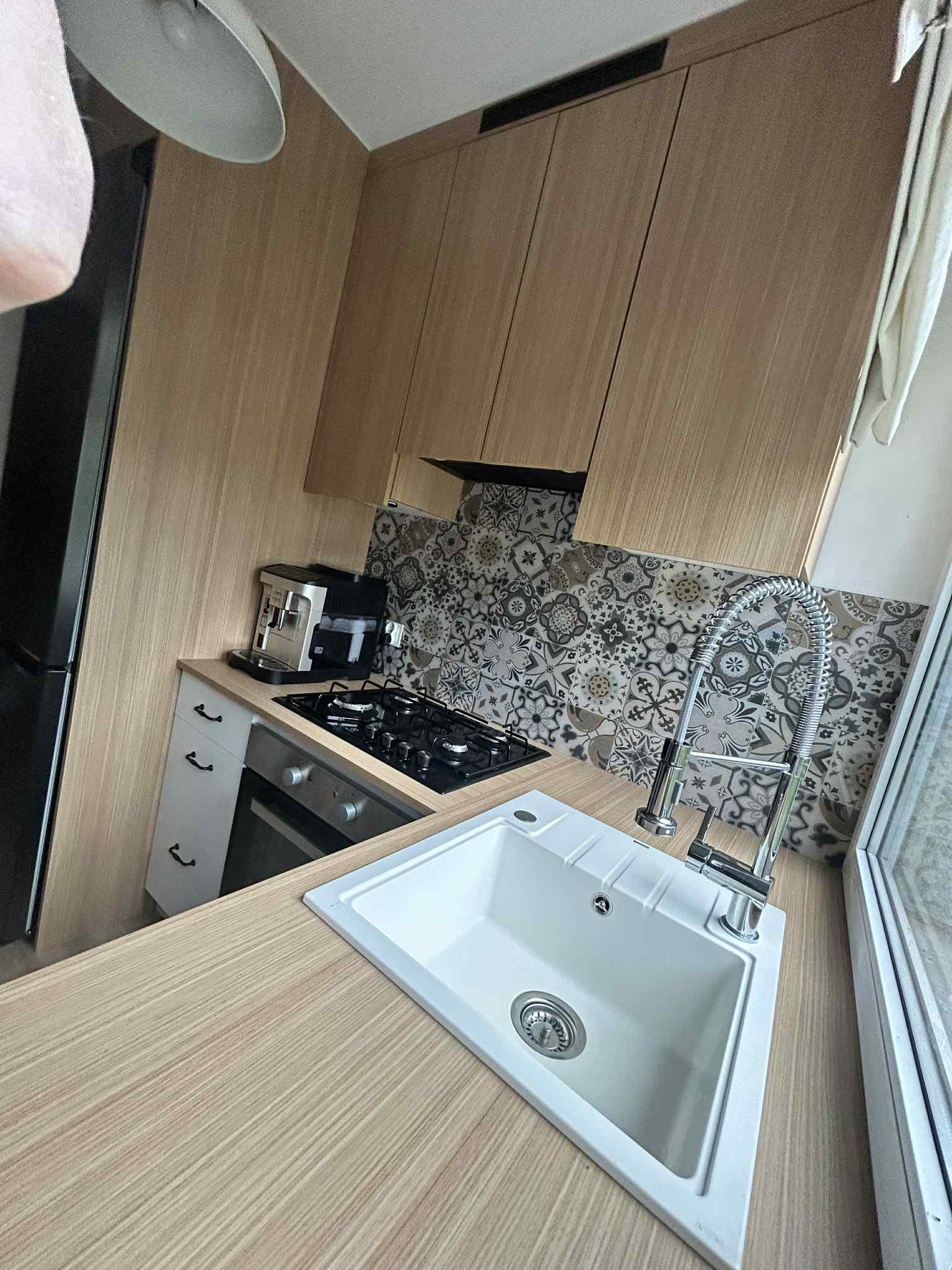 Jasna, zabudowana kuchnia z drewnianymi szafkami, wzorzystą ścianą nad blatem, zlewem i baterią. Widoczny ekspres do kawy i płyta gazowa. Ujęcie z perspektywy.