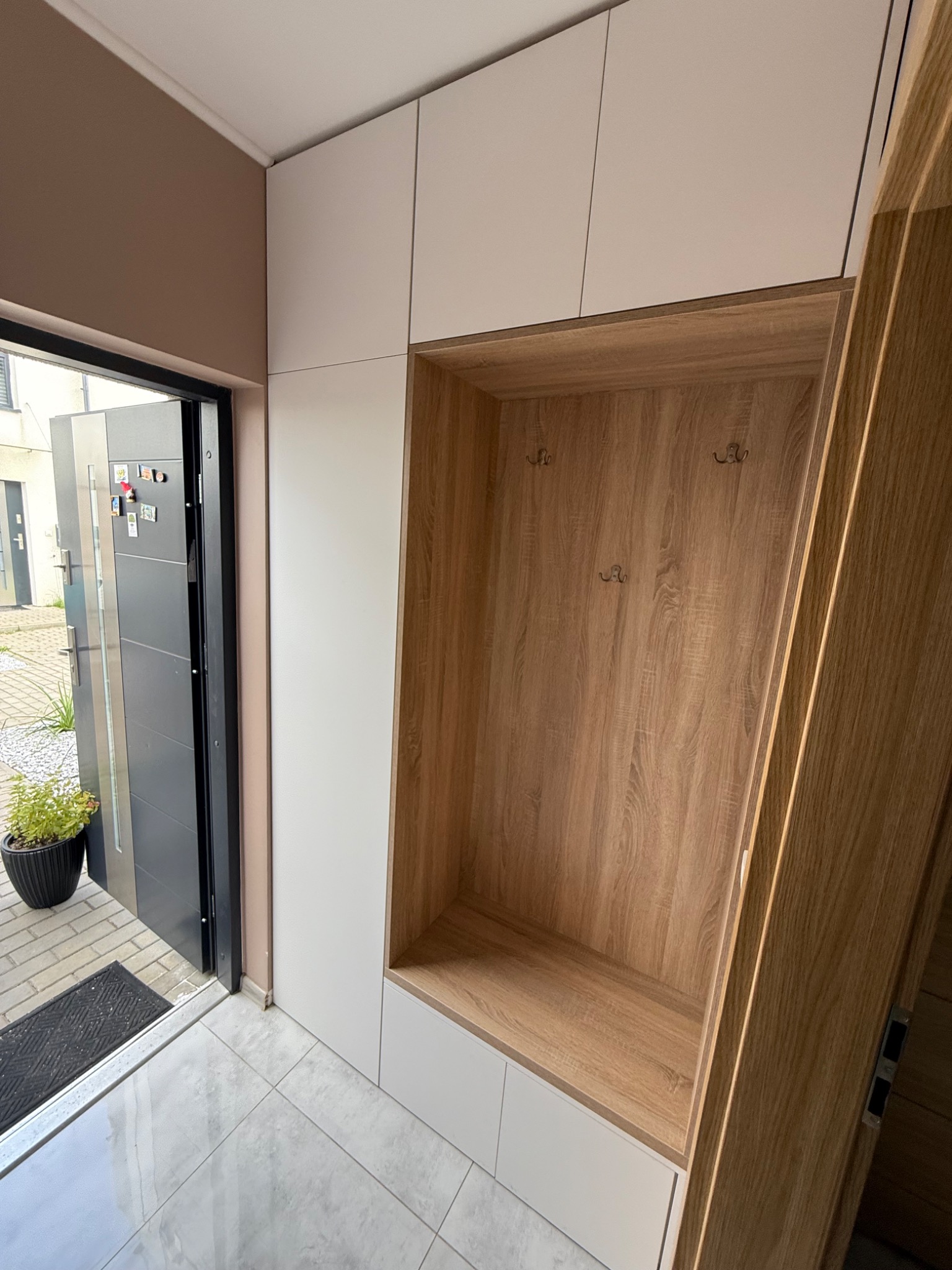 Funkcjonalna szafa wnękowa z wieszakami, drewnopodobne wykończenie, białe fronty, nowoczesny design w przedpokoju, widok na otwarte drzwi wejściowe.