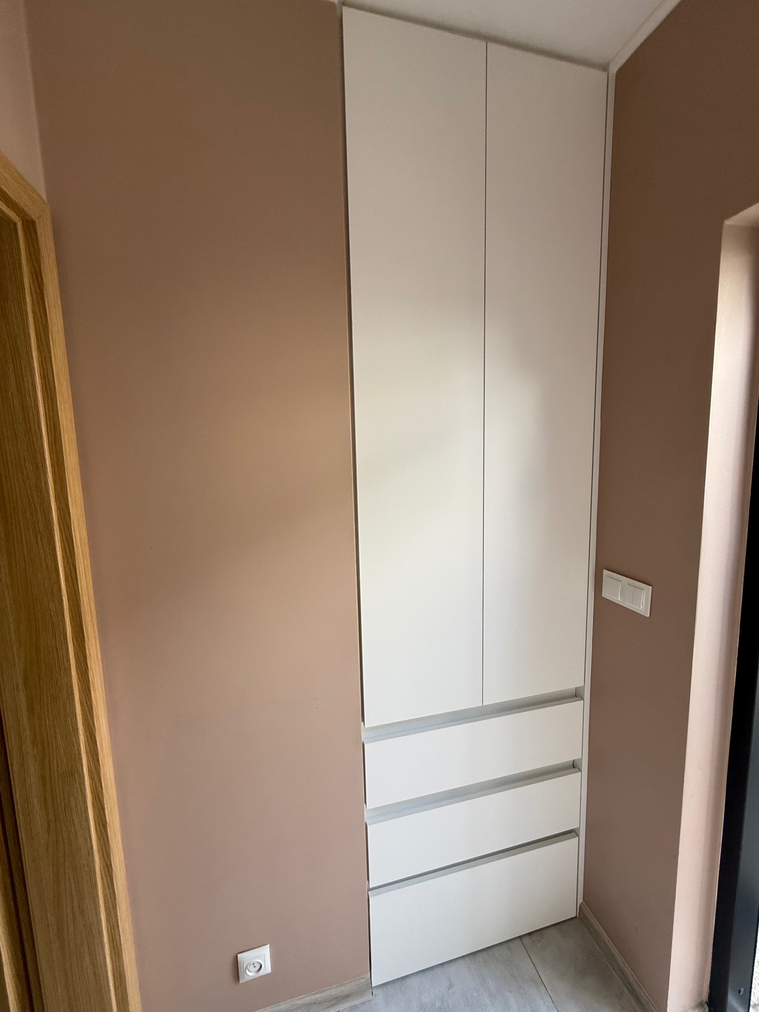 Biała szafa wnękowa z szufladami, dopasowana do ściany w odcieniach beżu, z widocznym gniazdkiem elektrycznym i fragmentem drzwi z ościeżnicą.