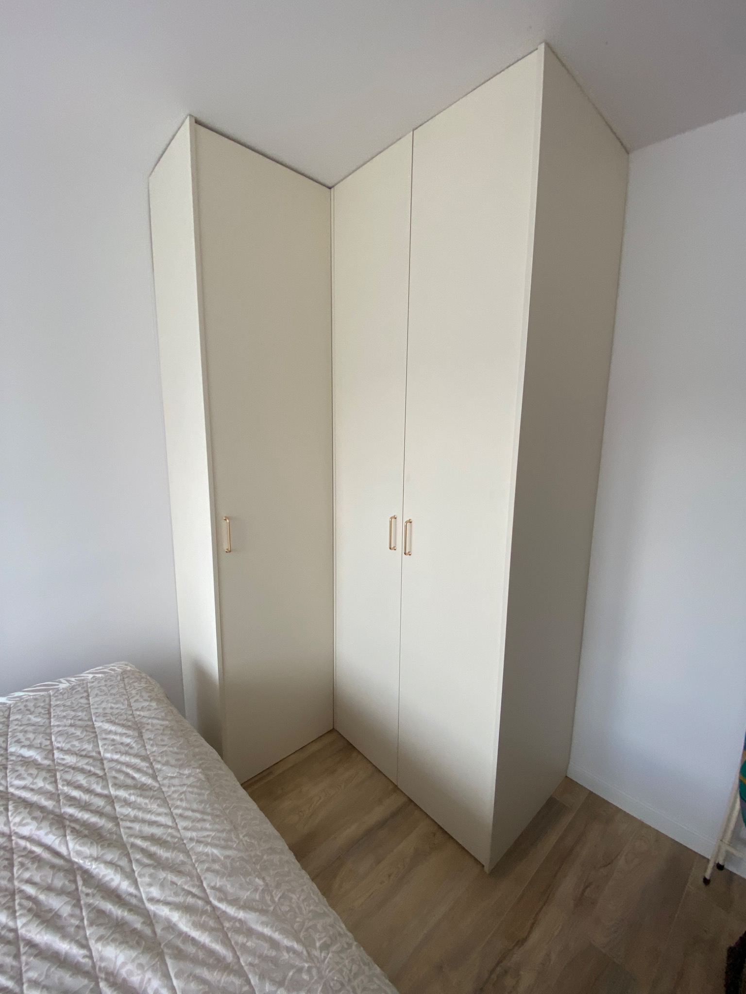 Biała, narożna szafa wnękowa z delikatnymi, złotymi uchwytami, dopasowana do skosu sufitu, widoczna z perspektywy łóżka w minimalistycznej sypialni.
