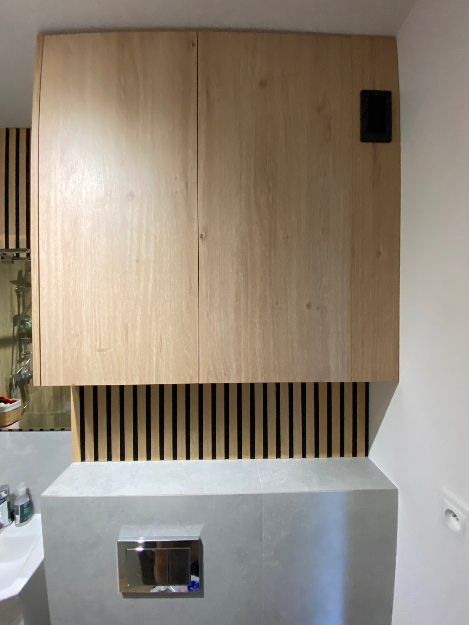 Drewniana szafka łazienkowa z lamelami w tle, nad zabudowaną toaletą. Minimalistyczny design, jasne kolory, widoczny fragment umywalki.