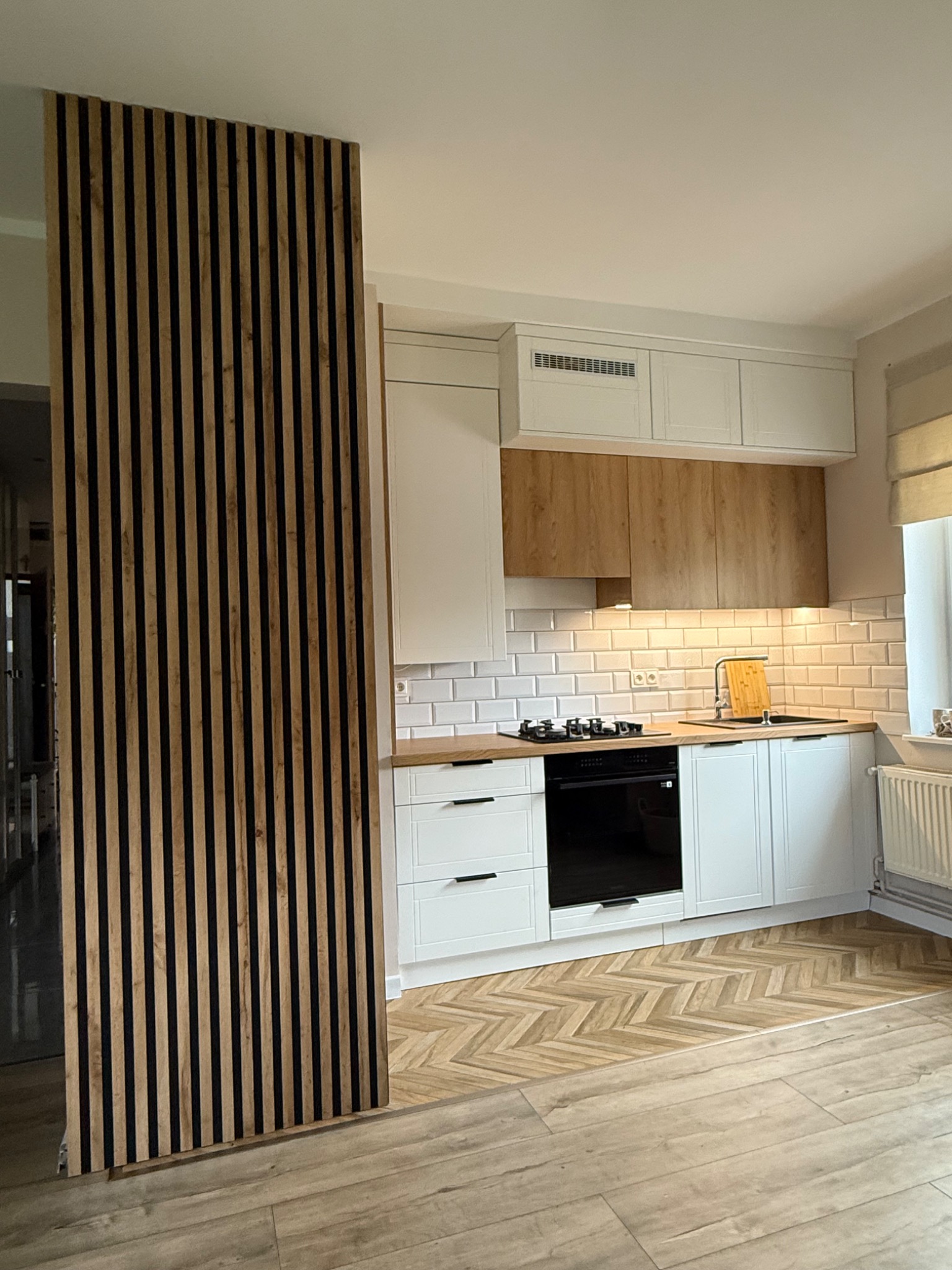 Nowoczesna kuchnia z białymi szafkami, drewnianym blatem i dekoracyjną ścianką z drewnianych listew. Parkiet ułożony w jodełkę. Ujęcie z perspektywy podłogi.