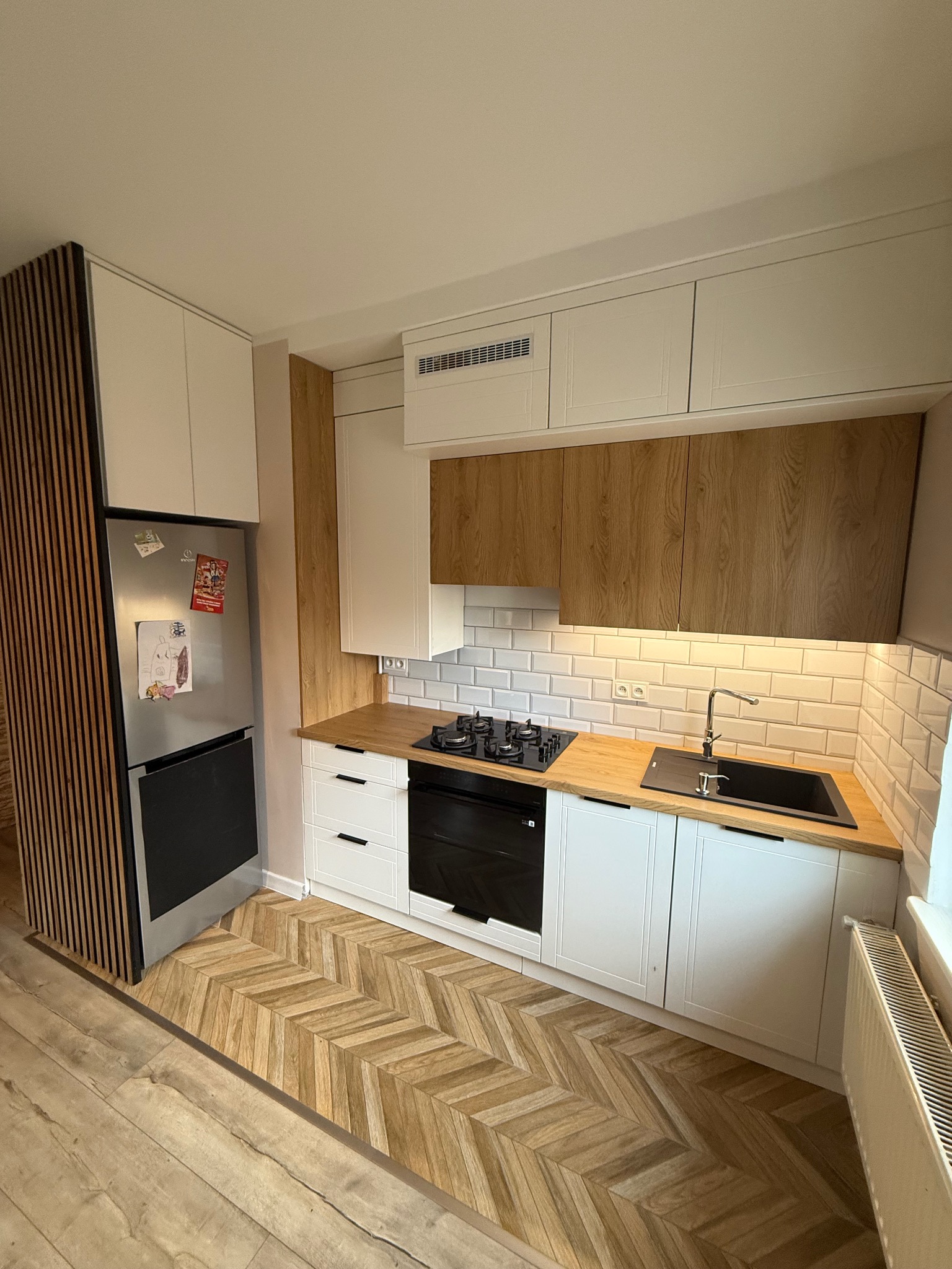 Nowoczesna kuchnia z drewnianymi akcentami: białe szafki, drewniany blat i podłoga w jodełkę. Lodówka obudowana drewnianymi lamelami. Czarny zlew i płyta.