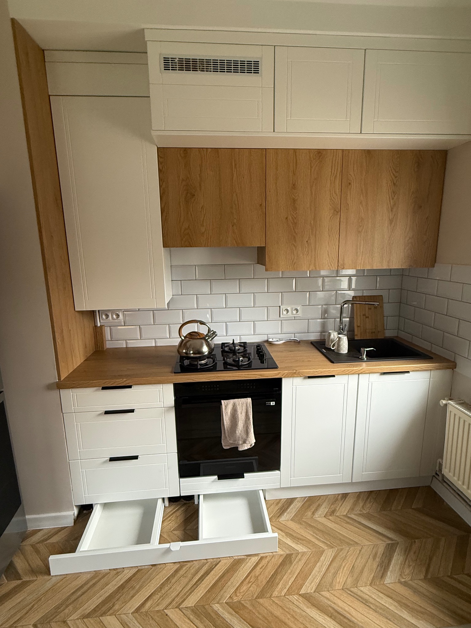 Nowoczesna kuchnia z białymi szafkami i drewnianymi akcentami. Otwarta szuflada dolna, parkiet w jodełkę, czarny zlew i płyta grzewcza. Minimalistyczny design.