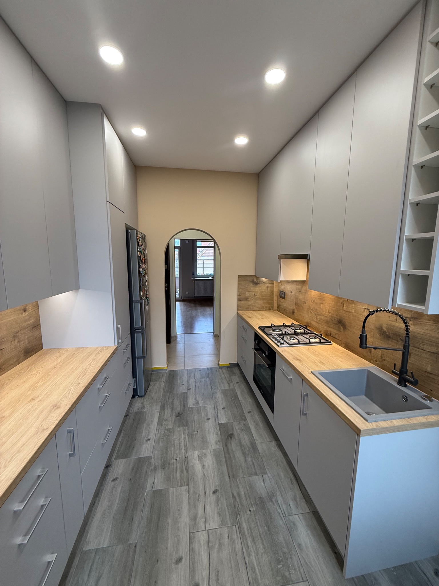 Wąska kuchnia w szarościach z drewnianymi blatami i płytkami podłogowymi imitującymi drewno. Widok na dalsze pomieszczenia przez łukowate przejście.