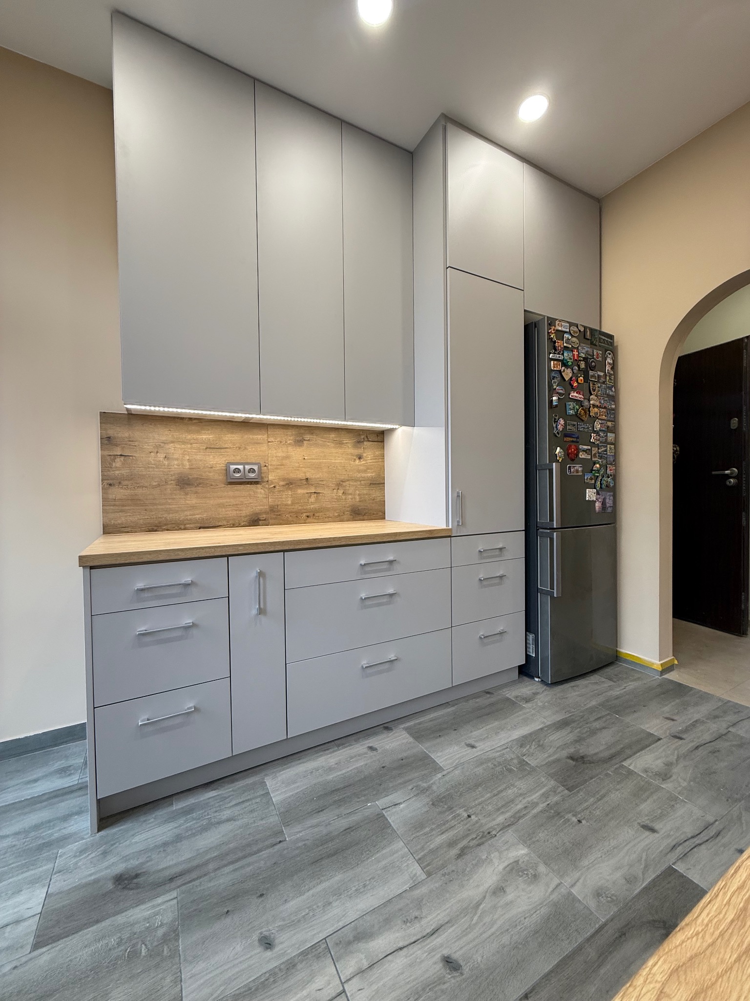 Szara zabudowa kuchenna z drewnianym blatem i oświetleniem LED, uzupełniona o lodówkę z magnesami, na tle szarej podłogi imitującej drewno.