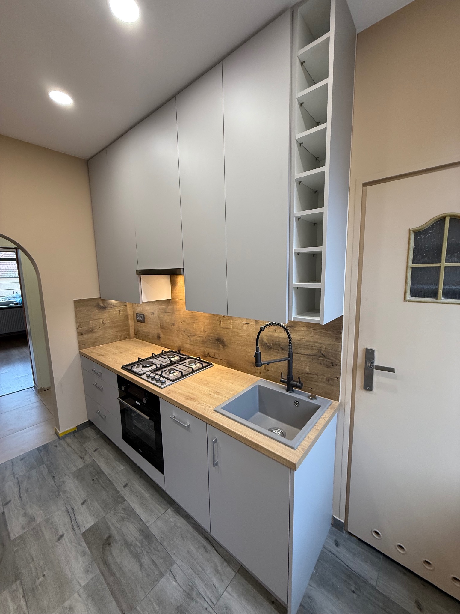 Szara kuchnia na wymiar z drewnianym blatem i płytkami imitującymi drewno na ścianie. Widoczny zlew, płyta gazowa i piekarnik. Nad blatem szafki wiszące.