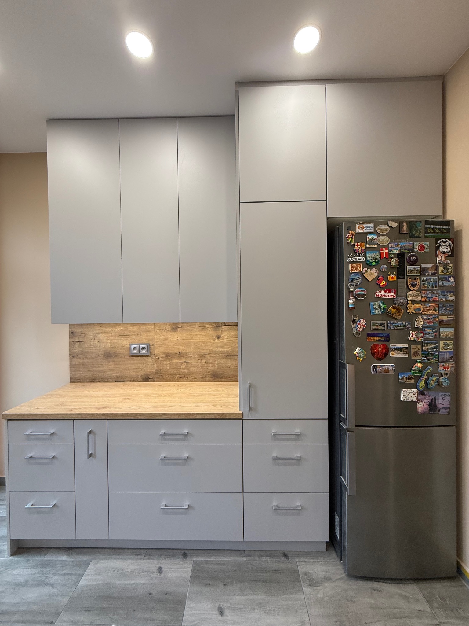 Jasnoszara kuchnia na wymiar z drewnianym blatem i lodówką obłożoną magnesami. Nowoczesny design, proste linie, funkcjonalne szafki i szuflady.