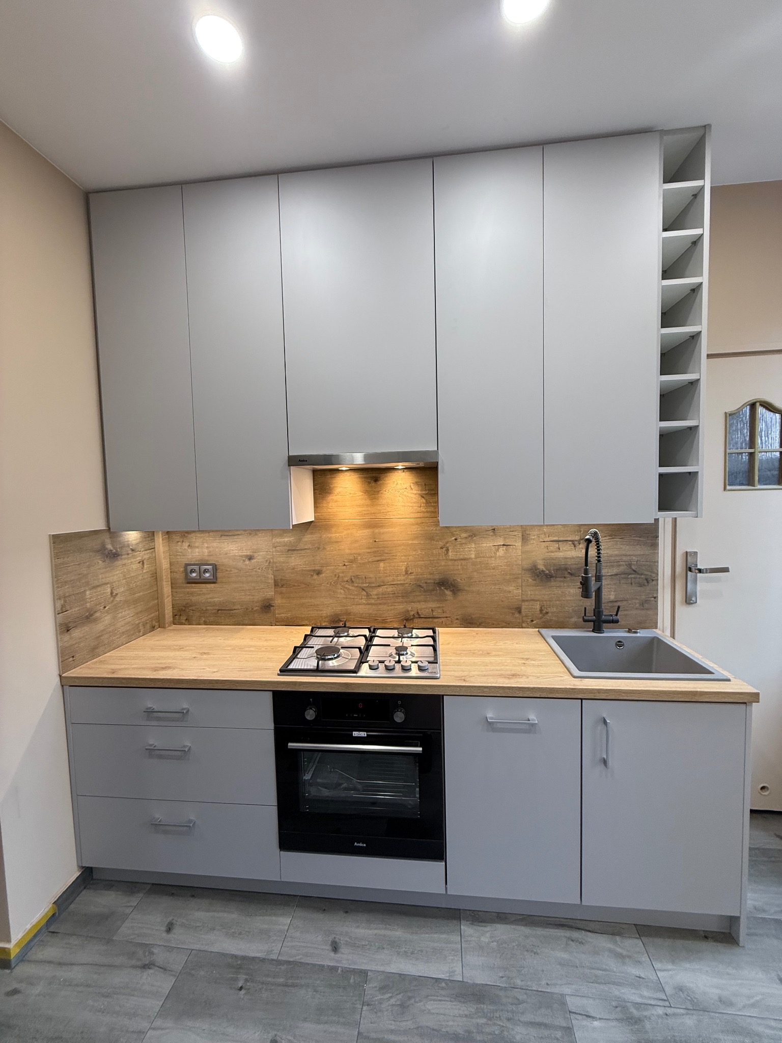 Szara kuchnia na wymiar z drewnianym blatem i płytkami imitującymi drewno na ścianie. Widoczna płyta gazowa, piekarnik i szara umywalka.
