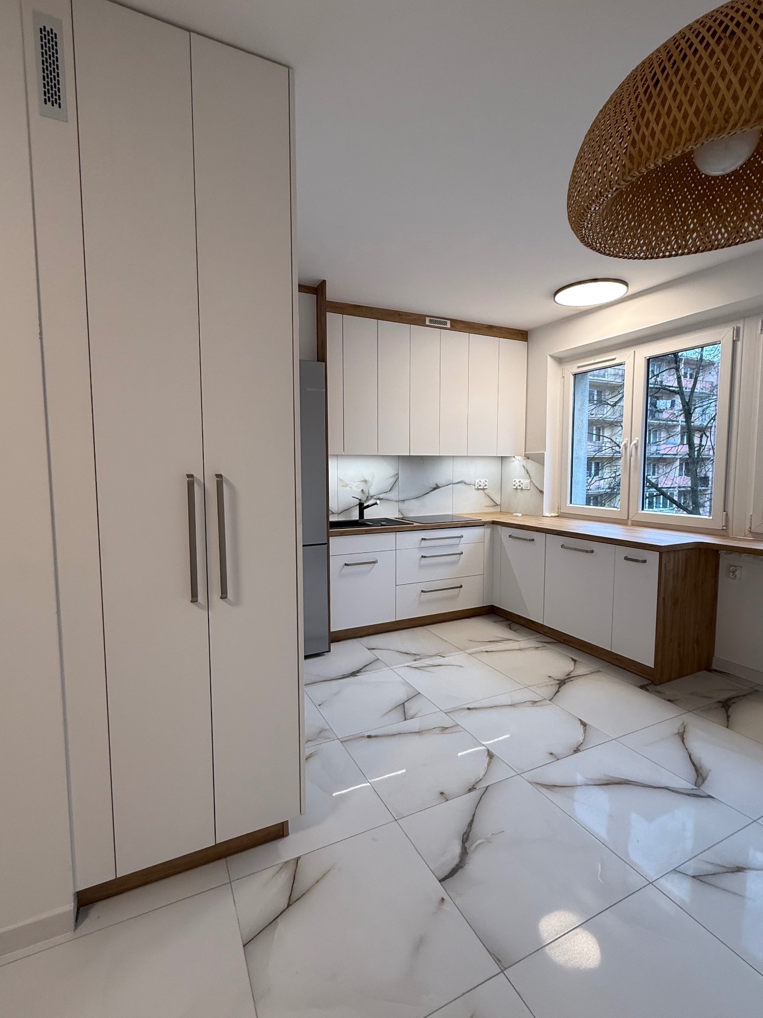 Nowoczesna kuchnia z białymi frontami i drewnianymi akcentami, marmurowa podłoga, widok na okno z ozdobną lampą. Minimalistyczny design z dbałością o detale.