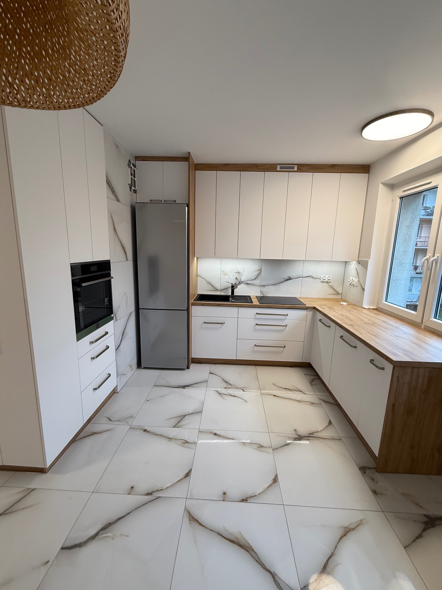 Nowoczesna kuchnia z białymi szafkami, drewnianym blatem i marmurową podłogą. Uwagę przykuwa designerska lampa wisząca i oświetlenie LED pod szafkami.