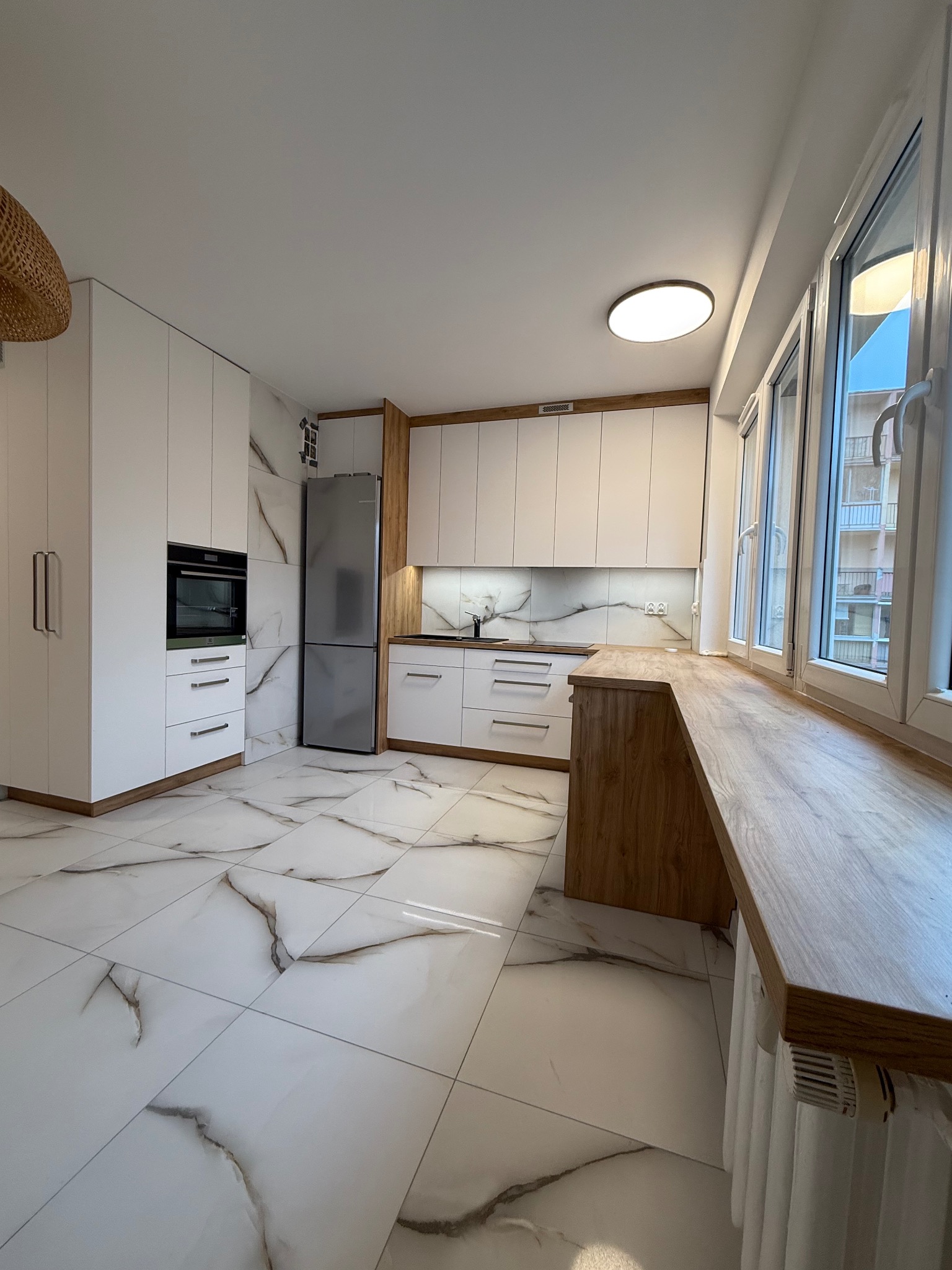 Nowoczesna kuchnia z białymi frontami i drewnianymi akcentami, marmurowa podłoga i blat przy oknie. Widok na zabudowę kuchenną z lodówką i piekarnikiem.